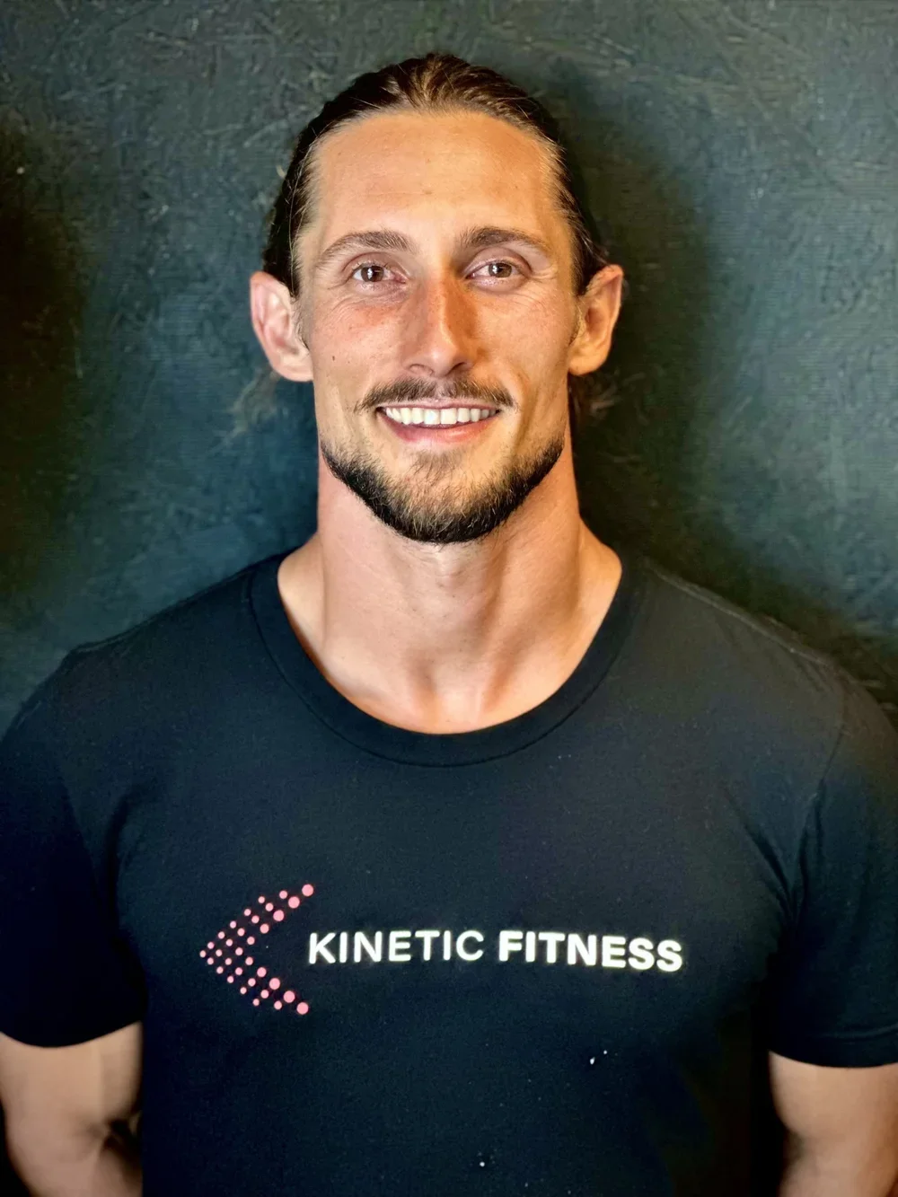 A smiling man with long hair tied back, a beard, and mustache, wearing a black T-shirt with the text "KINETIC FITNESS" and a pink dotted arrow logo, posed against a dark background.