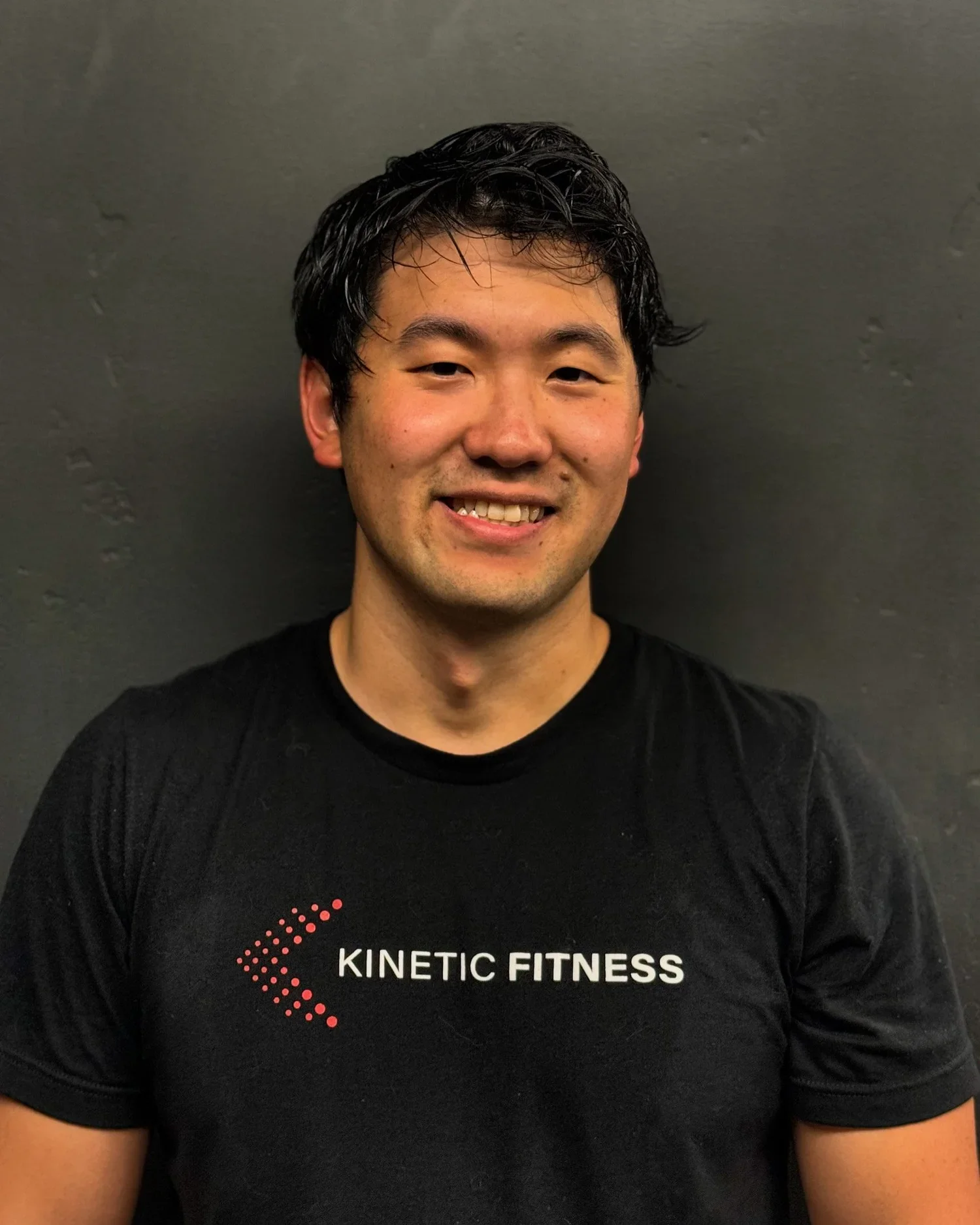 A smiling man wearing a black T-shirt with 'Kinetic Fitness' logo, standing against a dark gray wall.