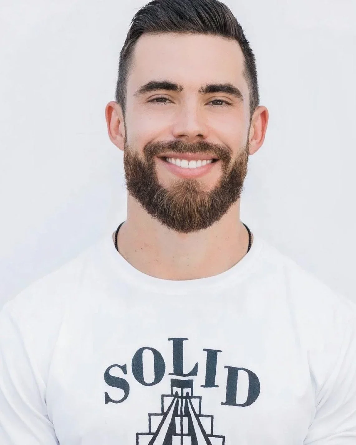 A smiling man with a beard and short dark hair, wearing a white T-shirt with a graphic and text, standing against a plain white background.