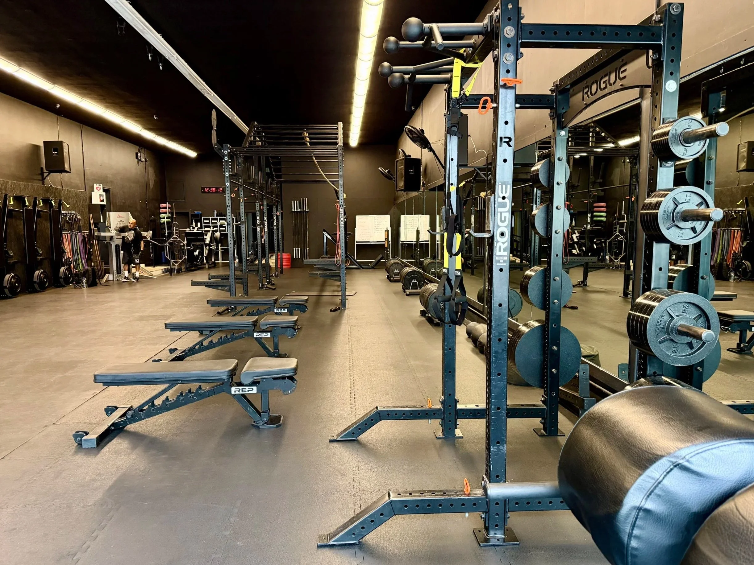 Inside a gym with black walls and flooring, various workout equipment including barbells, weight plates, and benches are visible. A person is seen in the distance, and a digital clock on the wall reads 12:38.