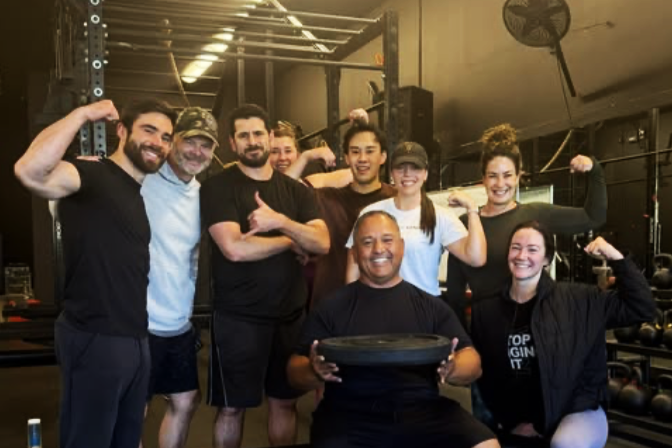 Group of nine fitness enthusiasts in gym, smiling and flexing muscles, with one person holding a weight plate.