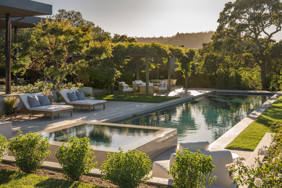 A luxurious backyard with a swimming pool, lounge chairs with cushions, and shaded seating areas surrounded by greenery at sunset.
