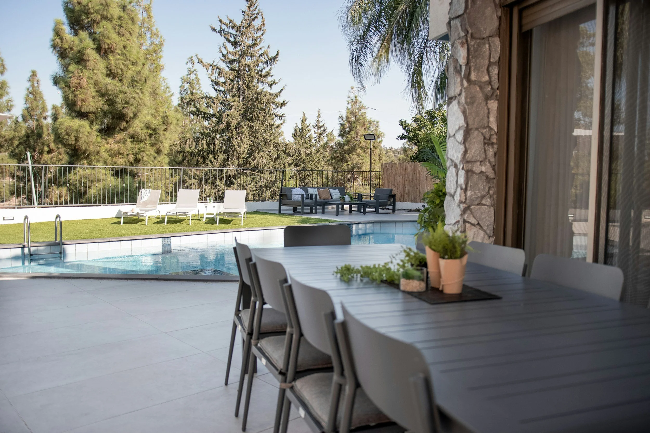 View of a backyard with a pool, white lounge chairs, a sitting area with a cushion, and a fence with tall pine trees in the background. The photo is taken from inside a covered patio area with a table, chairs, and potted plants.