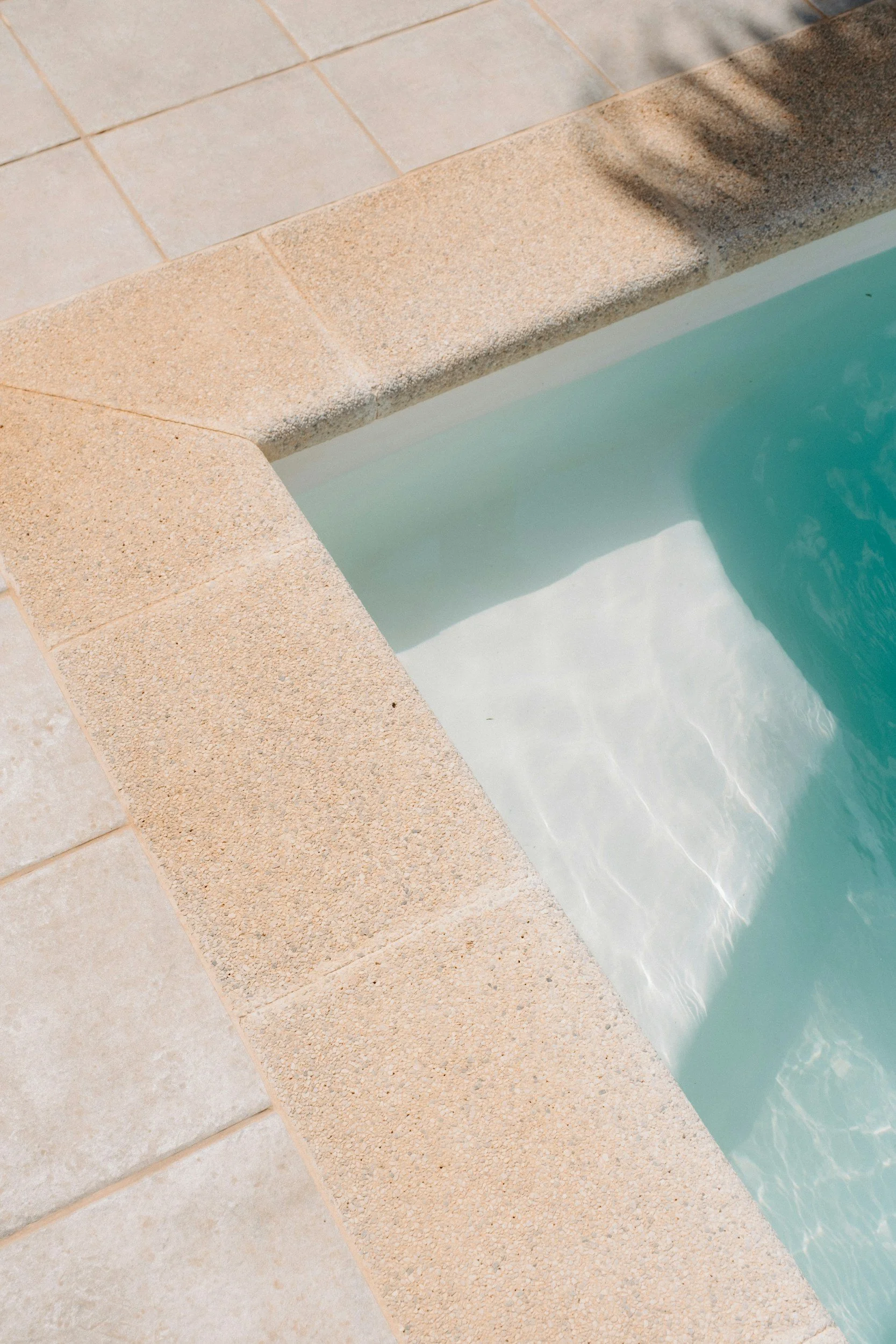 Close-up of a poolside with beige tiled border and a chain-link shadow cast on the edge. Clear turquoise water is visible in the pool.