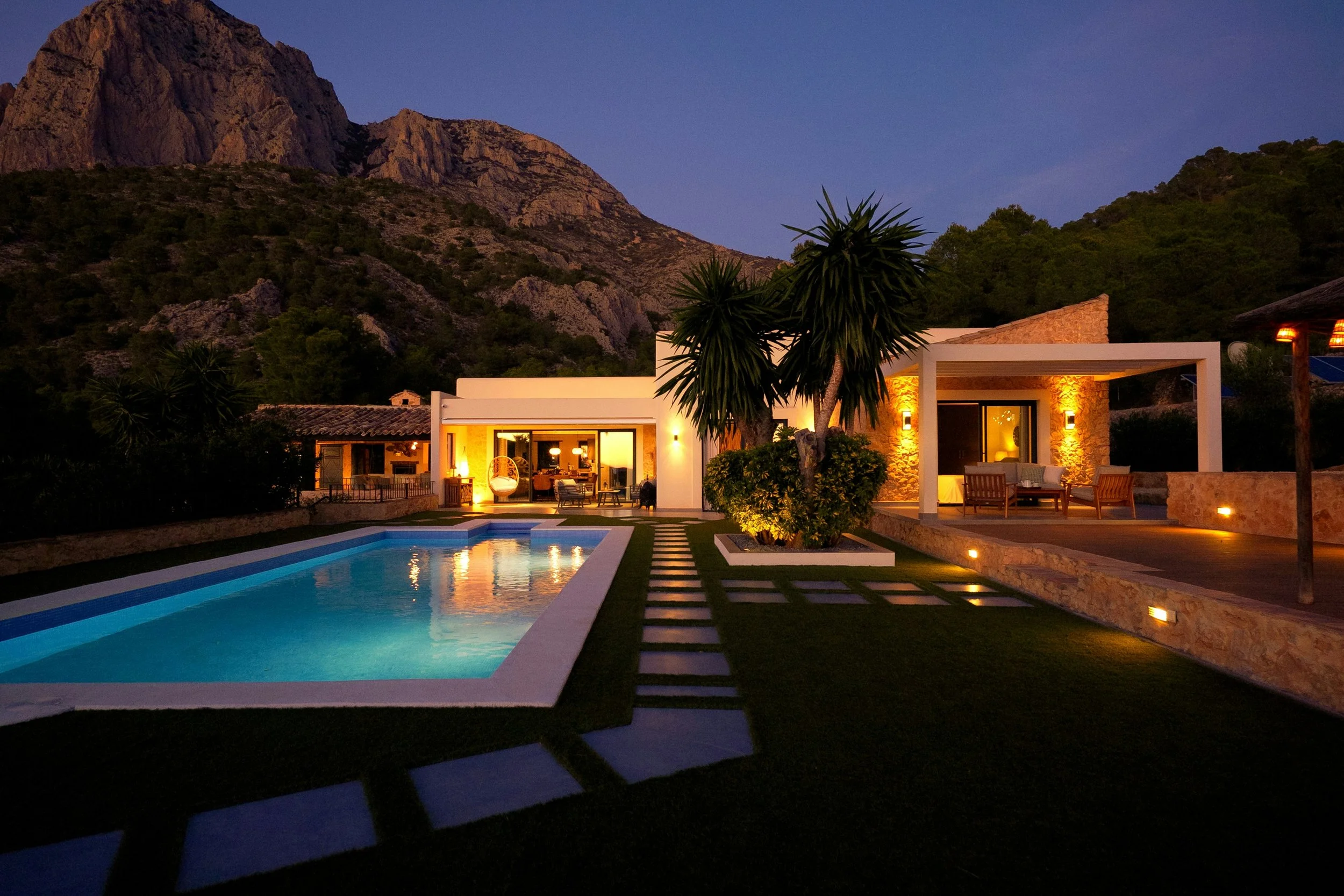 A modern house with large glass doors and a patio, illuminated by warm outdoor lighting, situated in a mountainous landscape during evening with a swimming pool in the foreground and trees nearby.