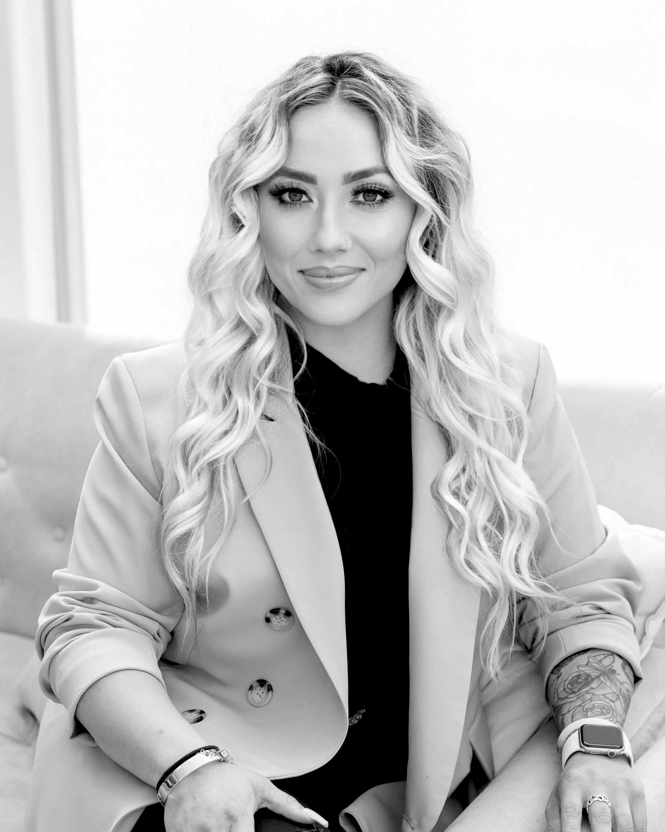 Black and white photograph of a woman with long, wavy blonde hair, smiling softly. She is wearing a light-colored blazer over a dark top, with makeup highlighting her eyes and lips. She has multiple bracelets on her left wrist, a ring on her right hand, and a tattoo of a floral design on her left forearm. She is sitting in front of a plain, light background.