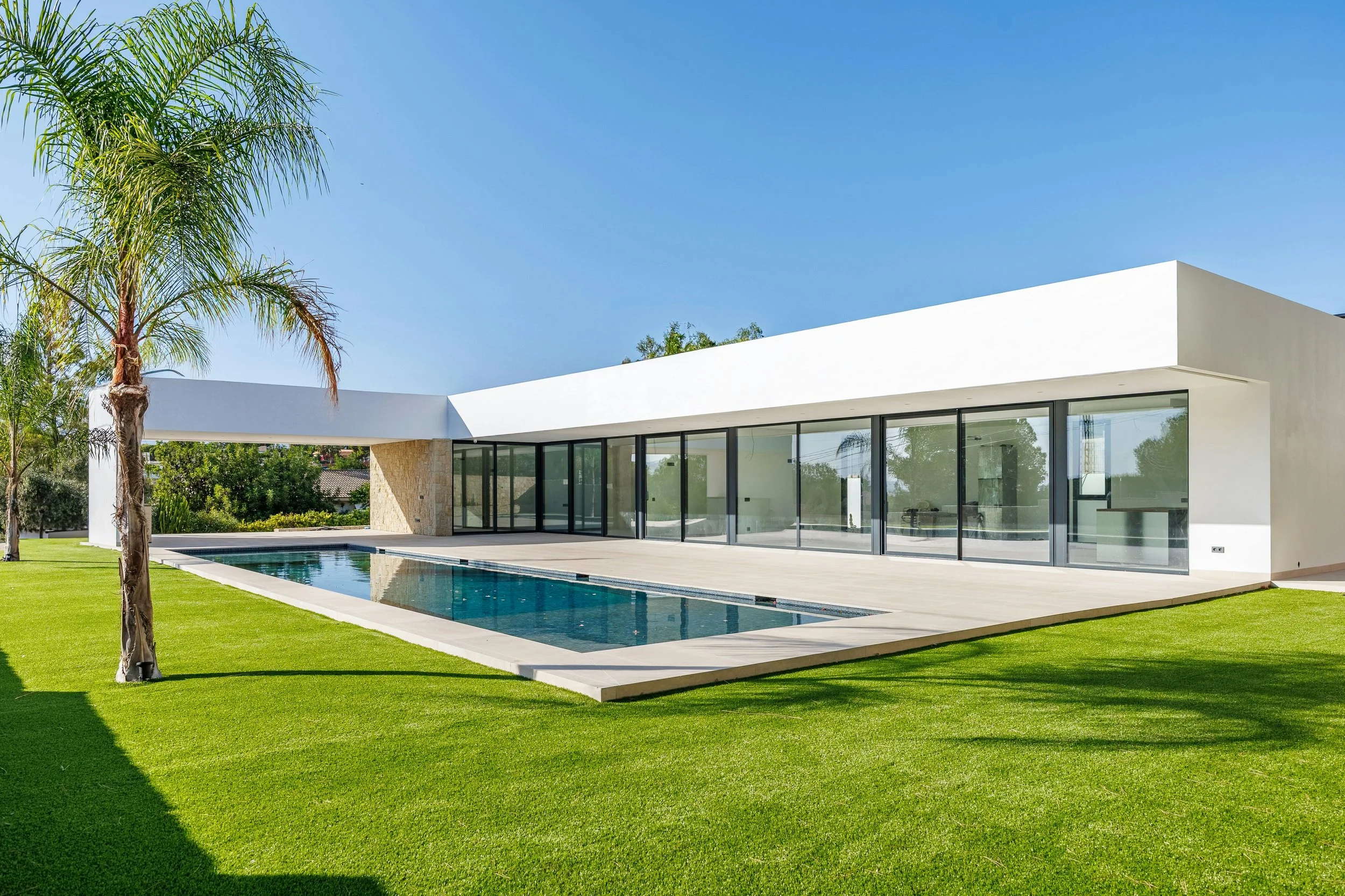 Modern white house with large glass windows and sliding doors, a swimming pool in the backyard, palm trees, and a well-maintained lawn under a clear blue sky.