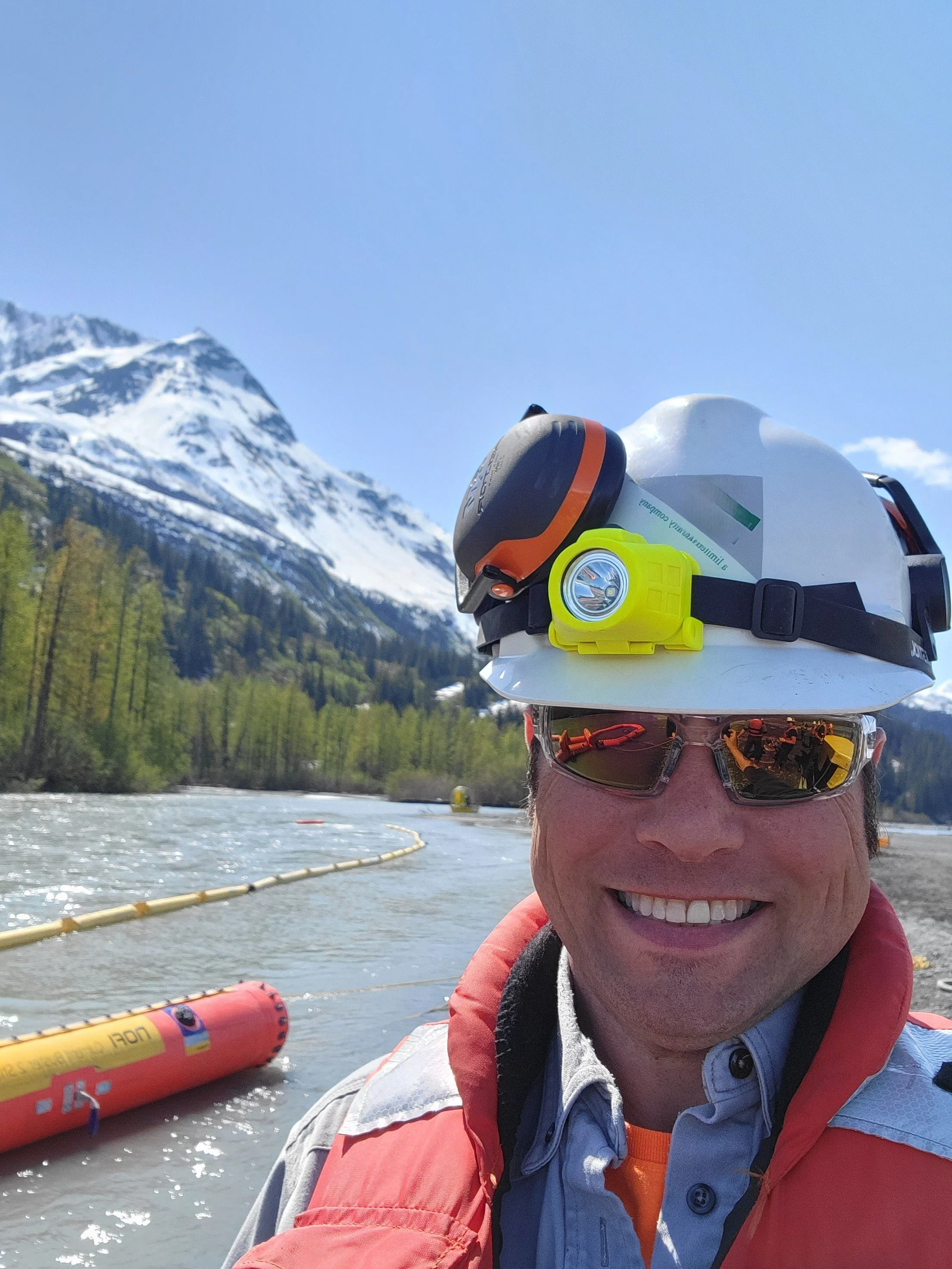 A smiling person wearing sunglasses, a white helmet with a headlamp, and a life jacket, standing near a body of water with snow-capped mountains in the background on a bright, sunny day.