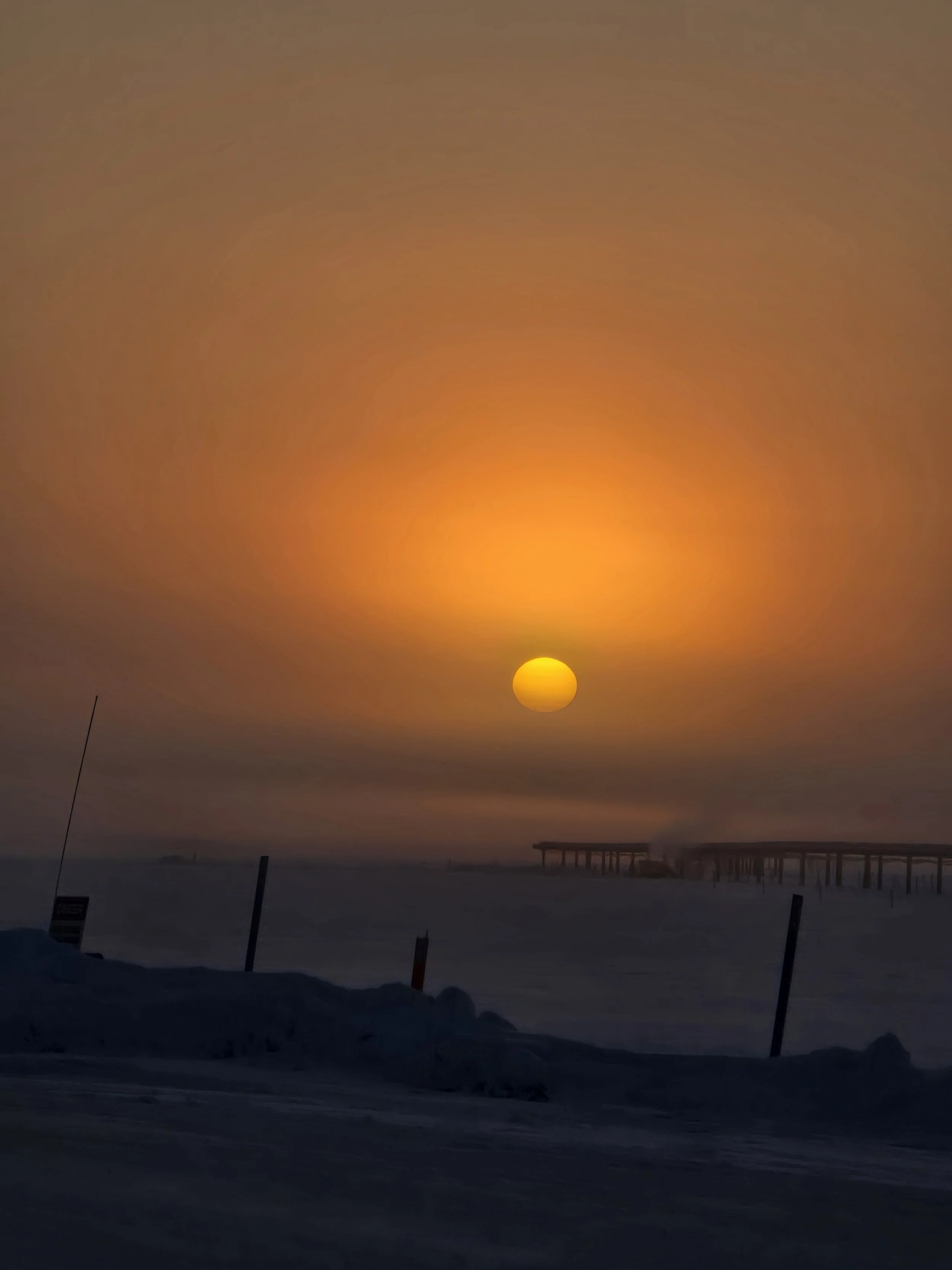 Sunset or sunrise over a snowy landscape with a pier extending into the water, dark sky, and silhouettes of snow-covered ground and dock structures.