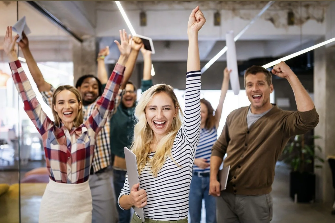 A group of diverse young professionals celebrating and cheering in an office setting, with their hands raised in victory and smiling happily.