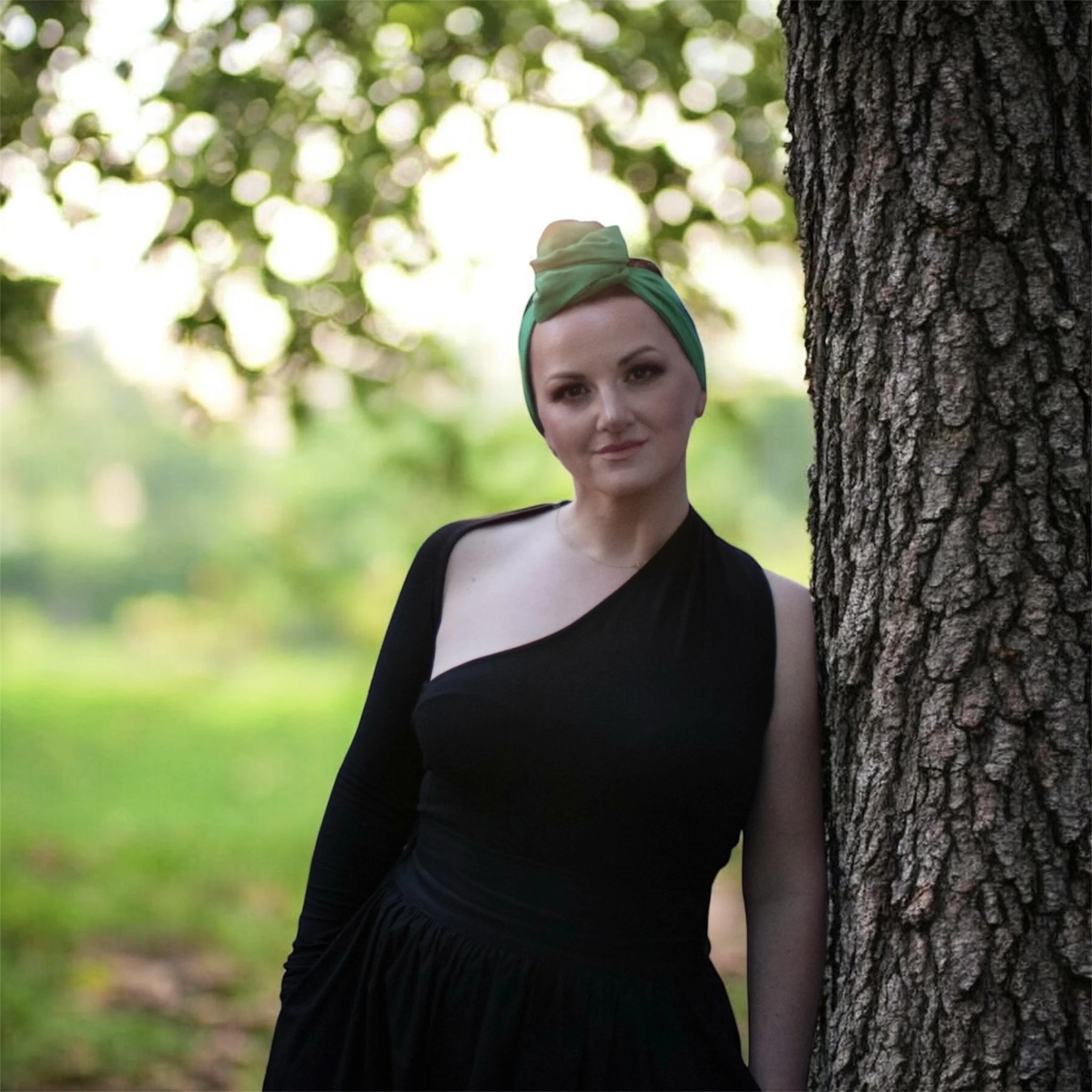 A woman with light skin and brown eyes standing outdoors, partially obscured by a large tree trunk with textured bark, against a background of green trees and blurred sunlight, wearing a green and yellow headscarf and black clothing.