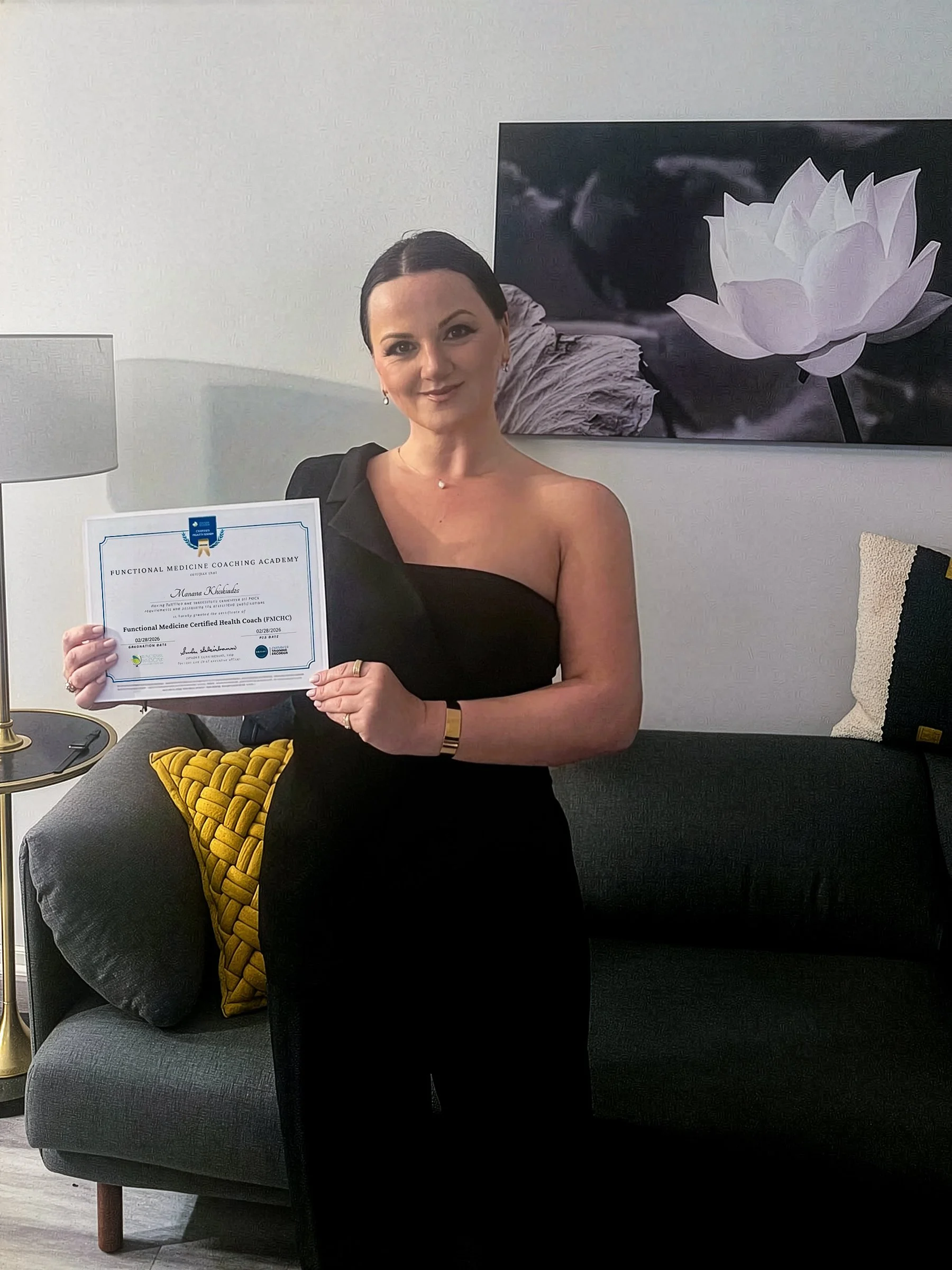 A woman in a black dress holding a certificate, seated on a gray sofa with a yellow pillow, in a room with white walls and modern decor.