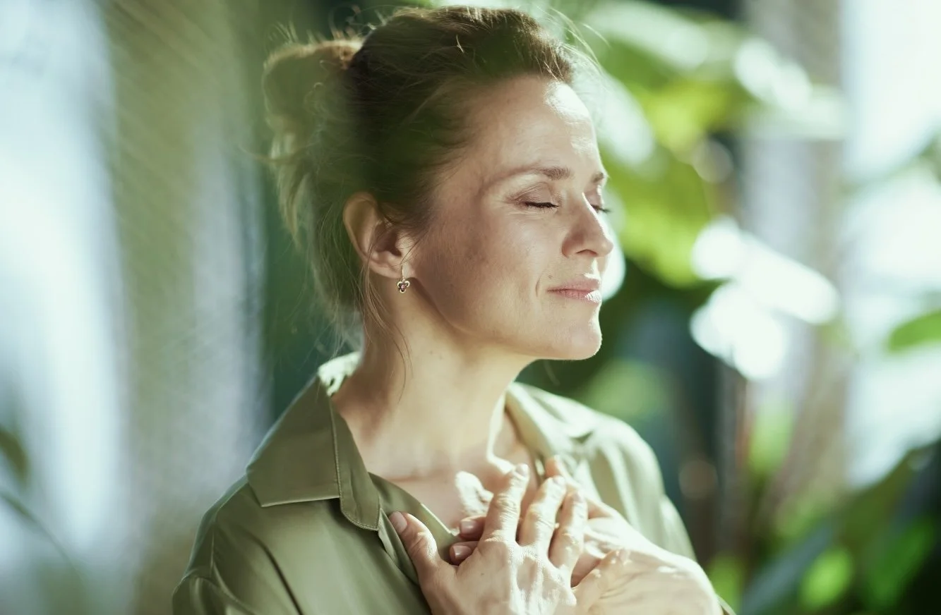 A woman with closed eyes and a peaceful expression, touching her chest with her hands, in soft natural light surrounded by blurred greenery.