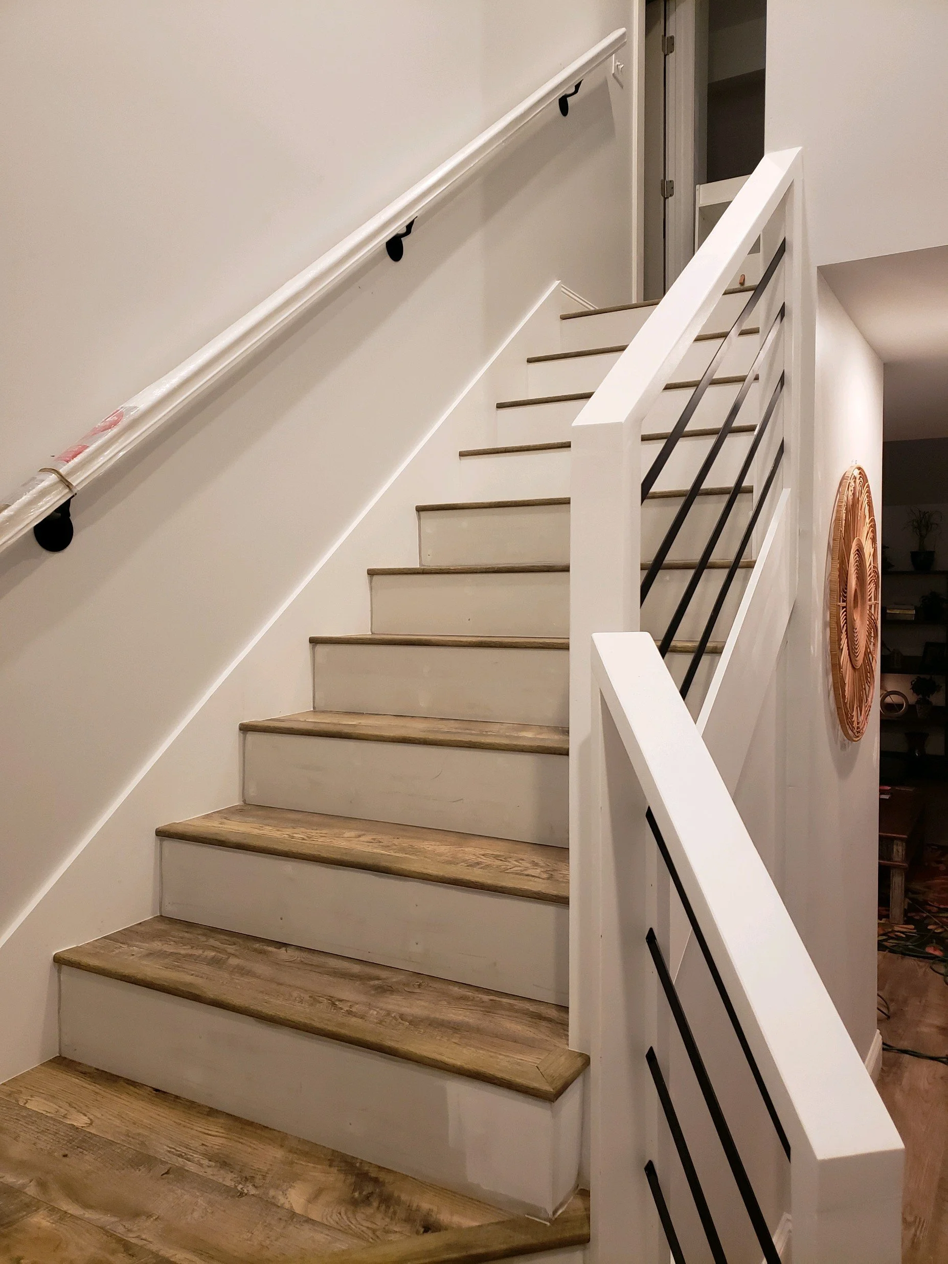 Interior view of a staircase with wooden steps, white risers, a white wall on the left side, and a white railing with black horizontal bars on the right side. A section of a decorative wall art or clock is visible on the right wall.
