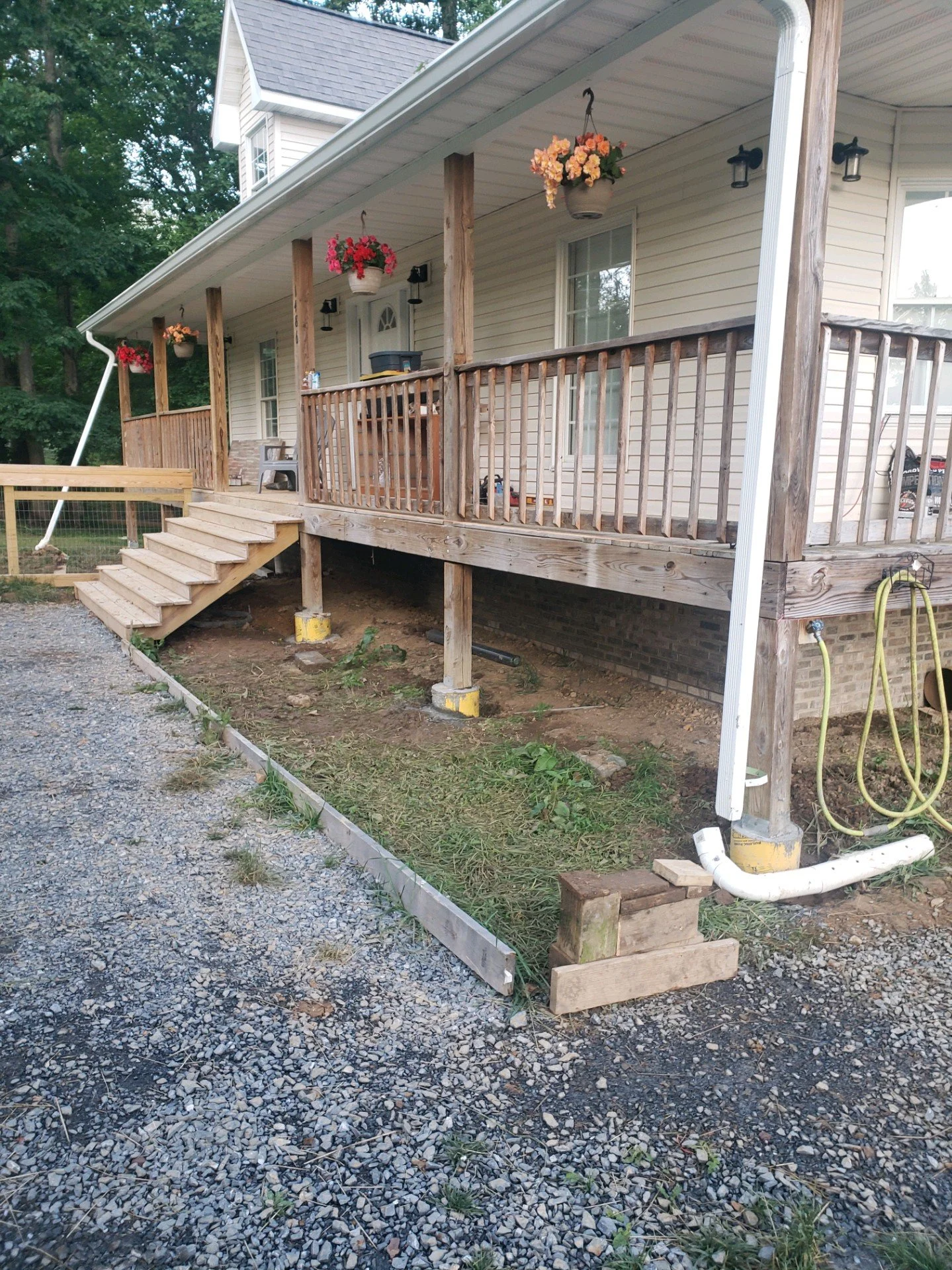 A house with a wooden deck under construction, with stairs leading up to the deck. There are hanging flower pots with colorful flowers, and various tools and materials around the deck area. The ground below is gravel with some patches of grass.