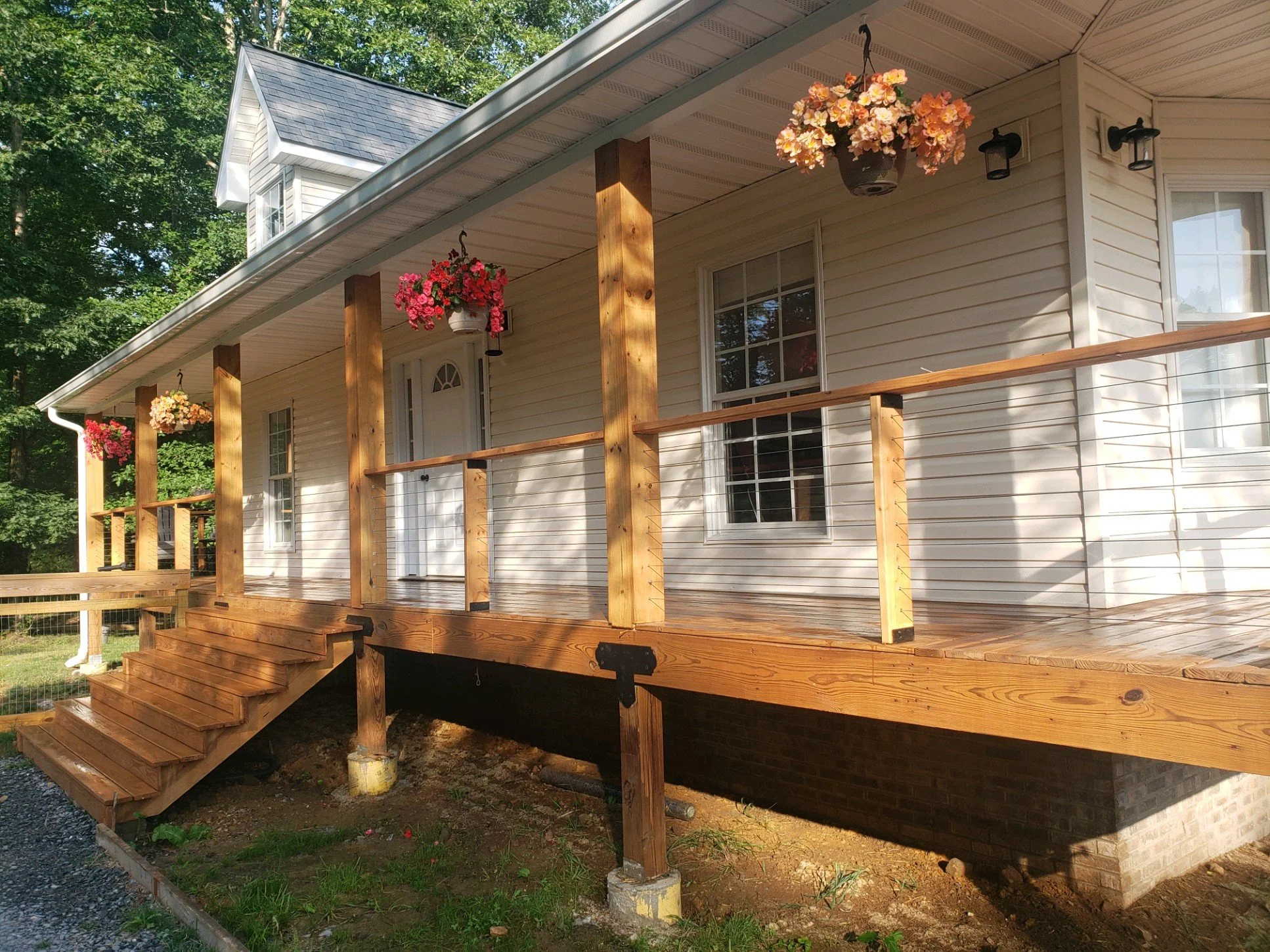 A house with a porch under construction, showing wooden staircase, support beams, hanging flower baskets, and exterior walls with siding.