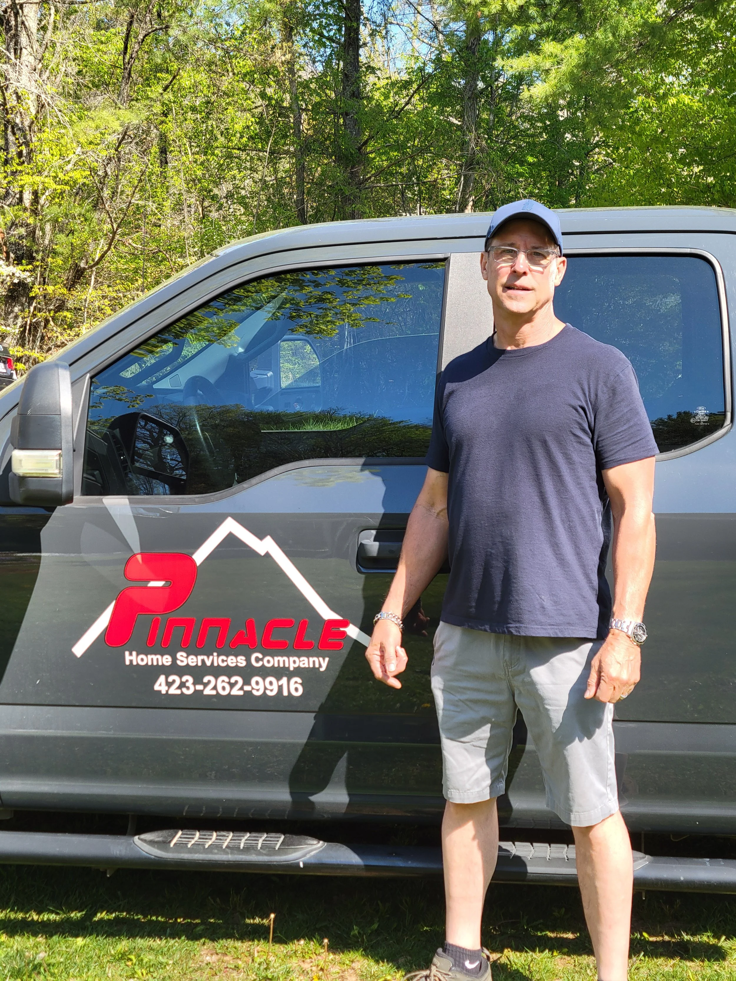A man wearing glasses, a white cap, a black t-shirt, and gray shorts standing outdoors next to a black service vehicle with a logo and contact information for a home services company. The background features green trees and blue sky.