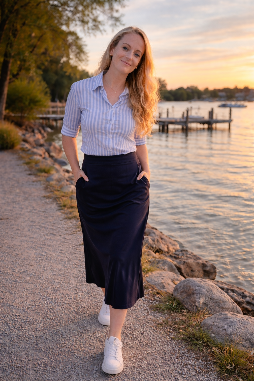 Eine junge Frau geht an einem Ufer bei Sonnenuntergang spazieren, trägt eine blaue gestreifte Bluse, einen schwarzen Rock, weiße Turnschuhe, hat lang gewellte blonde Haare und lächelt.