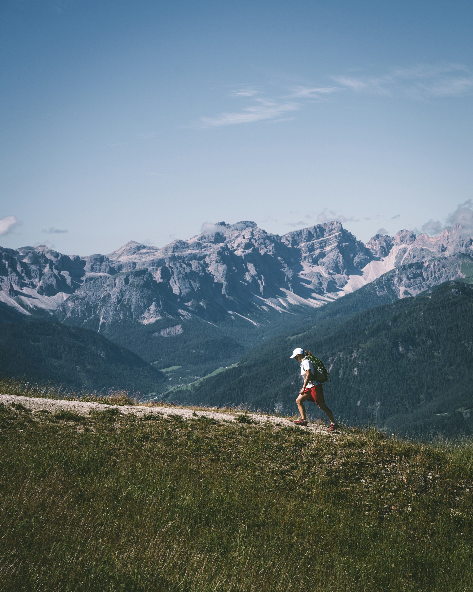 A person hiking along a mountain trail with a backdrop of a mountain range under a partly cloudy sky.