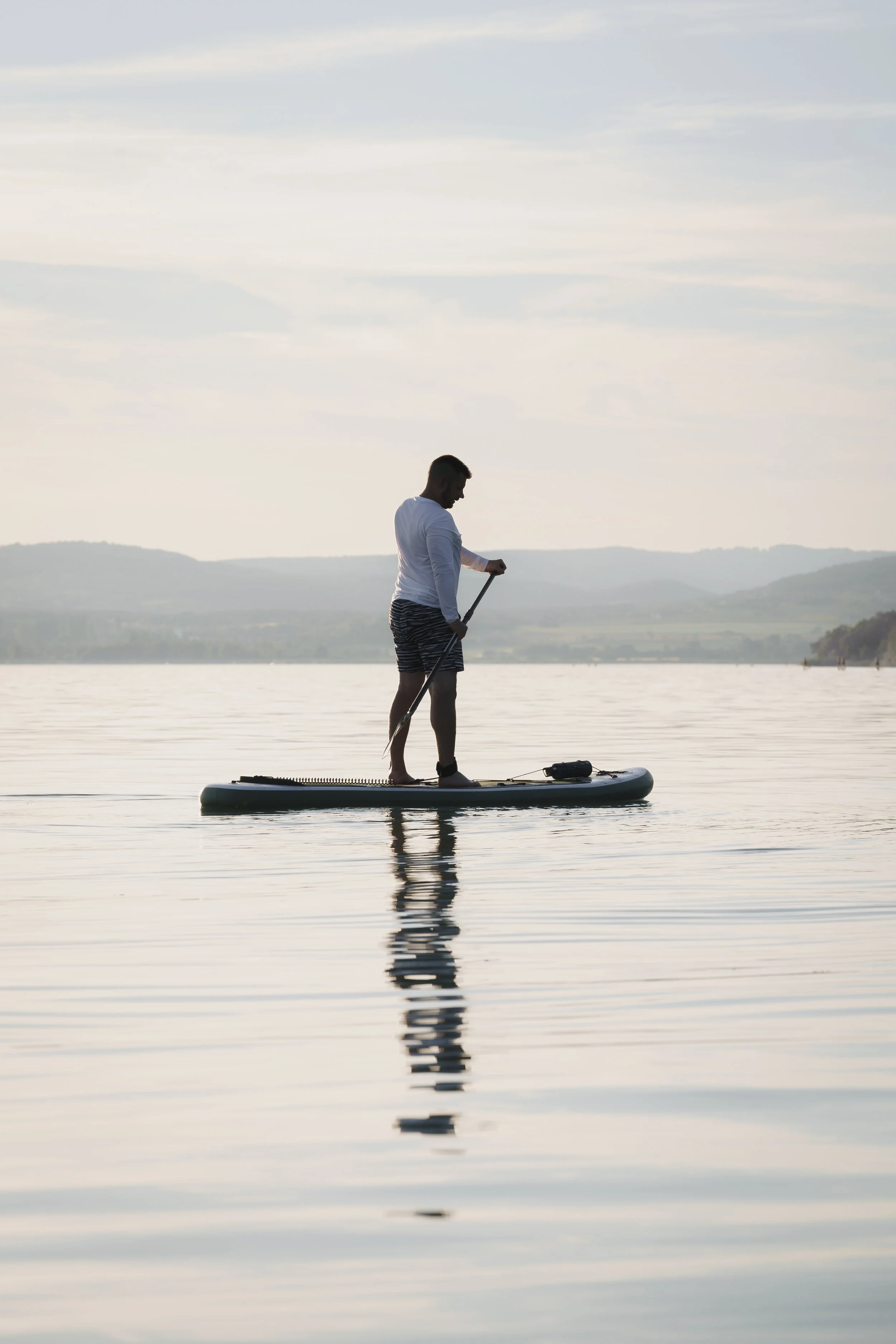 A man standing on a paddleboard on a calm lake, holding a paddle, with distant hills and a cloudy sky in the background.
