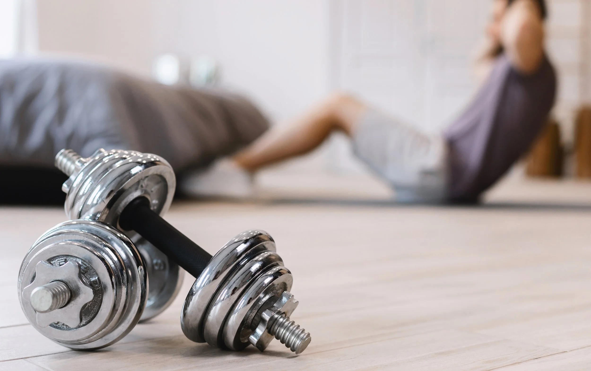 Close-up view of a metallic dumbbell placed on a light-colored wooden floor, with a blurred person in workout attire in the background.