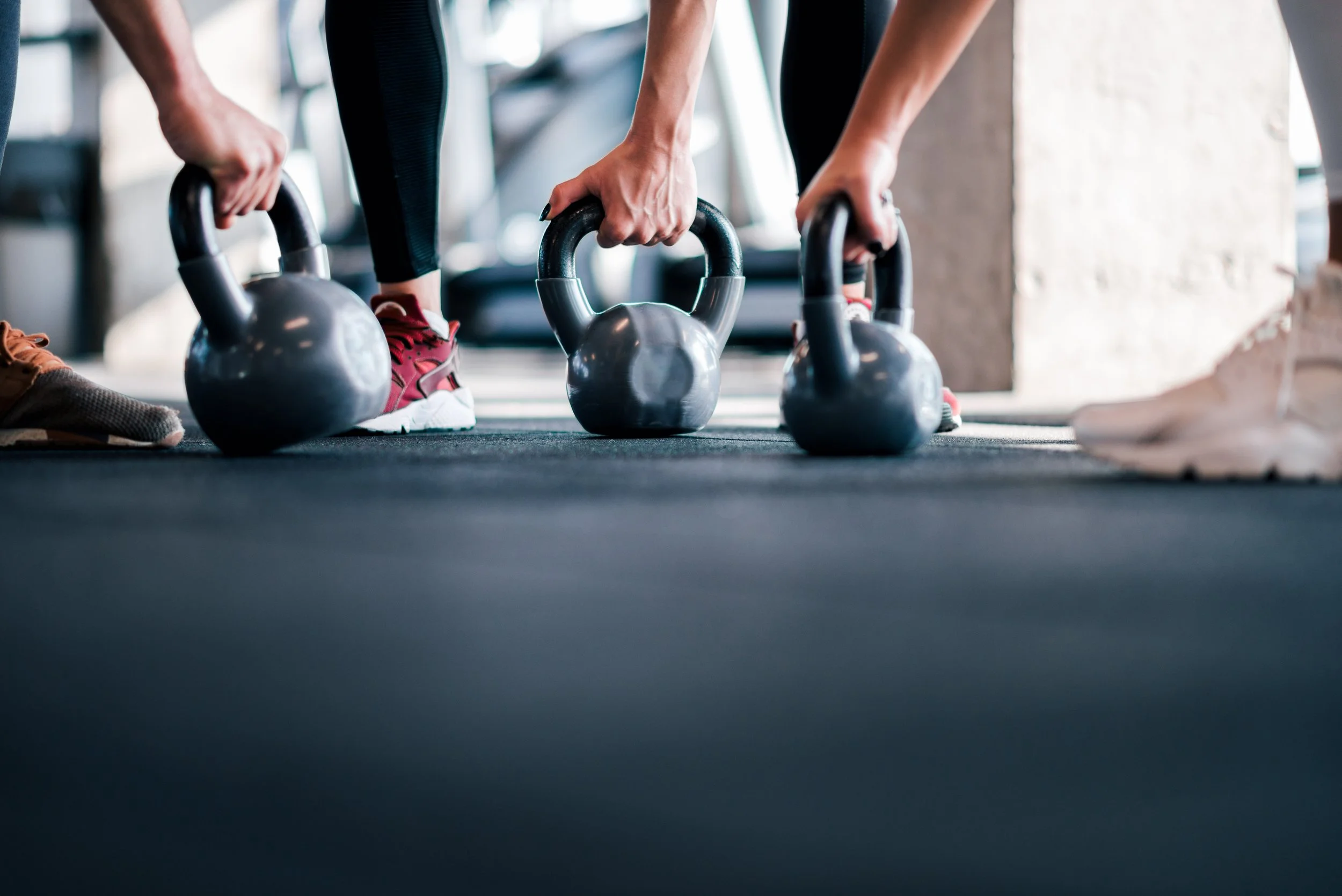 People performing a kettlebell workout in a gym, lifting kettlebells on a black mat.