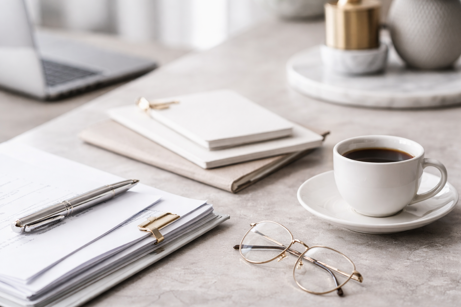 Desk workspace with a laptop, notebooks, a pen, glasses, a cup of coffee, and some decorative items.