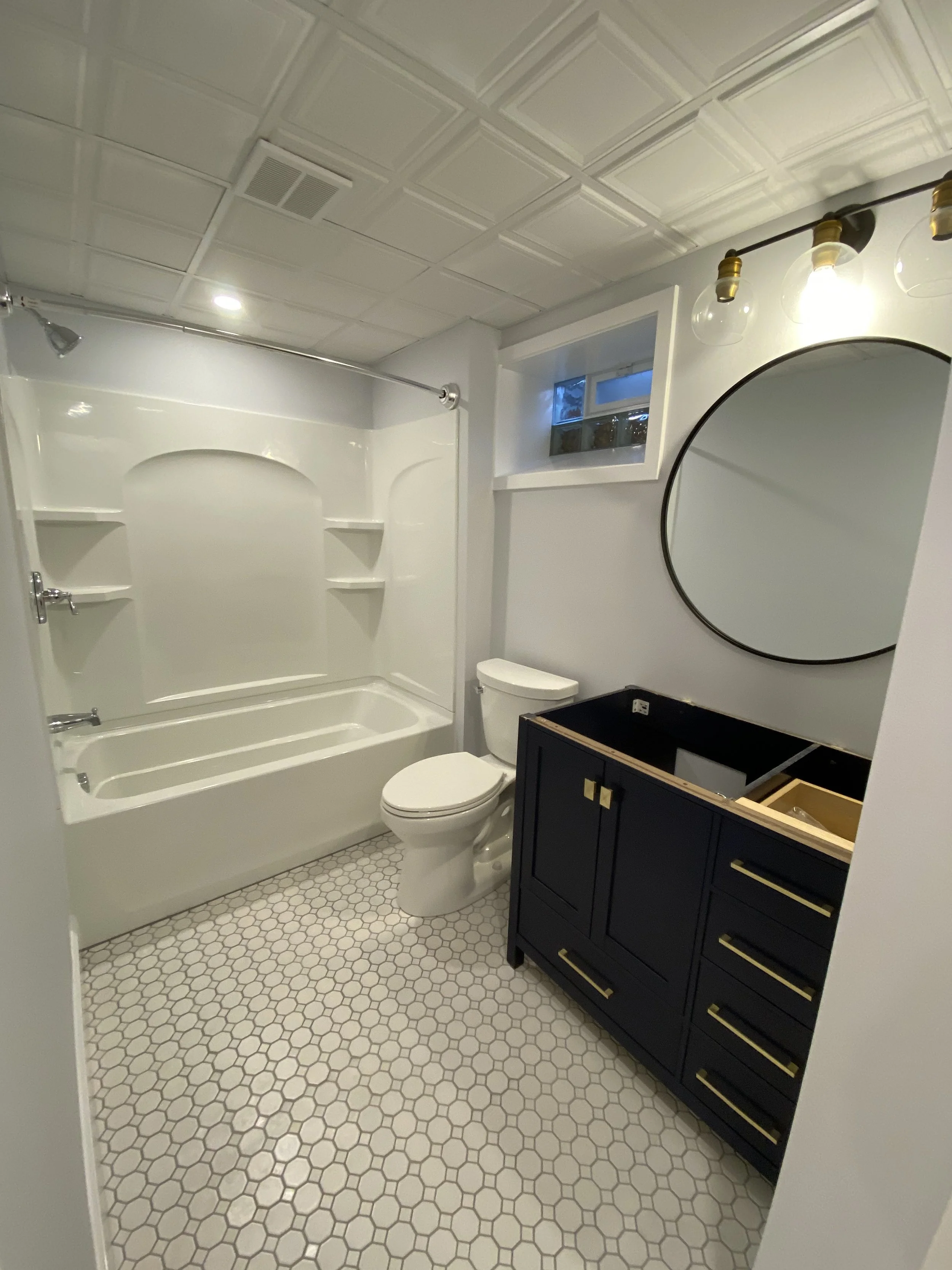 A bathroom with a white bathtub with shelves, a toilet, a navy blue vanity with gold handles, a round mirror, a black counter, a window, and a ceiling with decorative tiles.