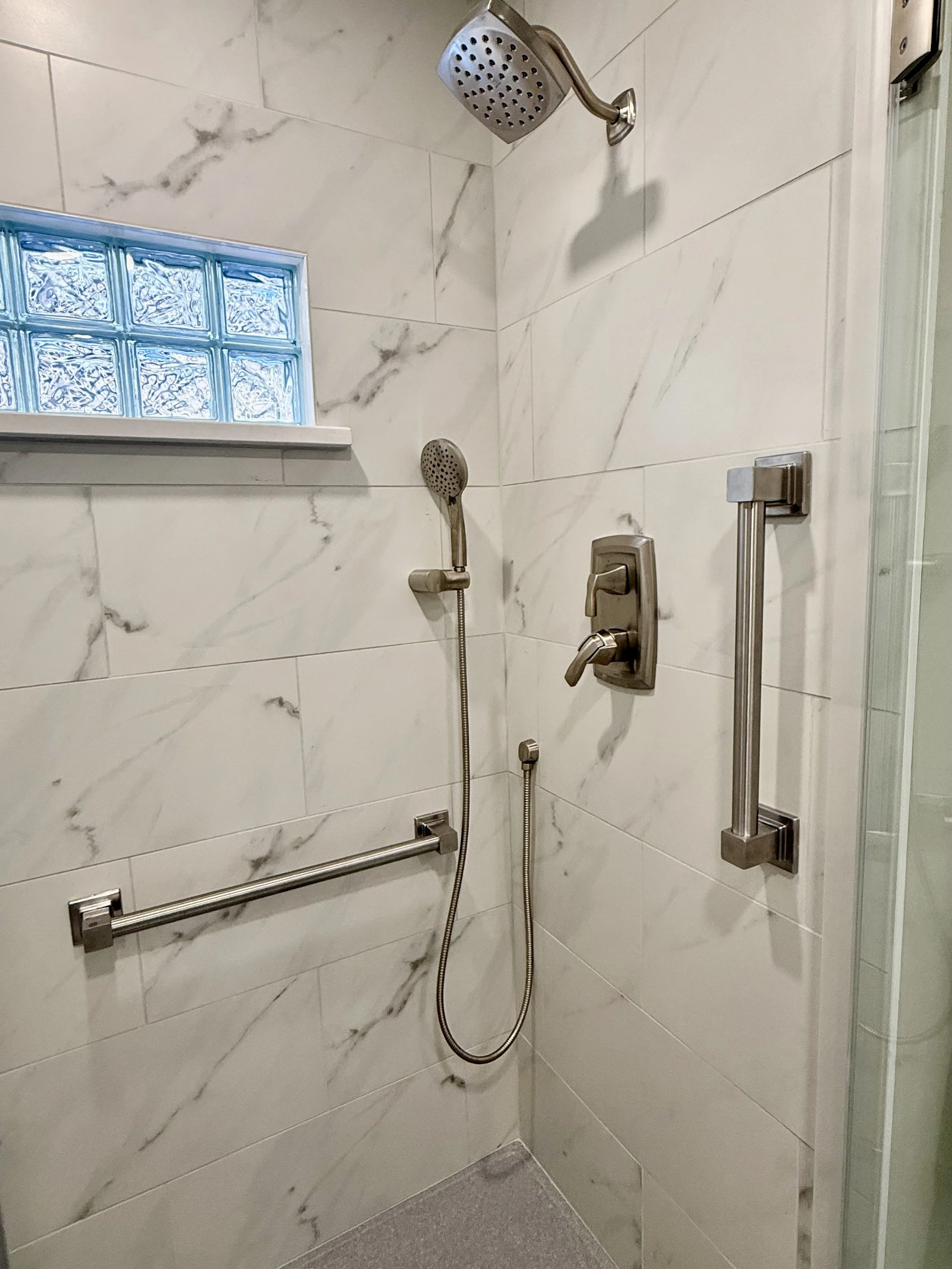 Shower with marble tiles, a shower head, a handheld shower spray, grab bars, and a small window with glass blocks.