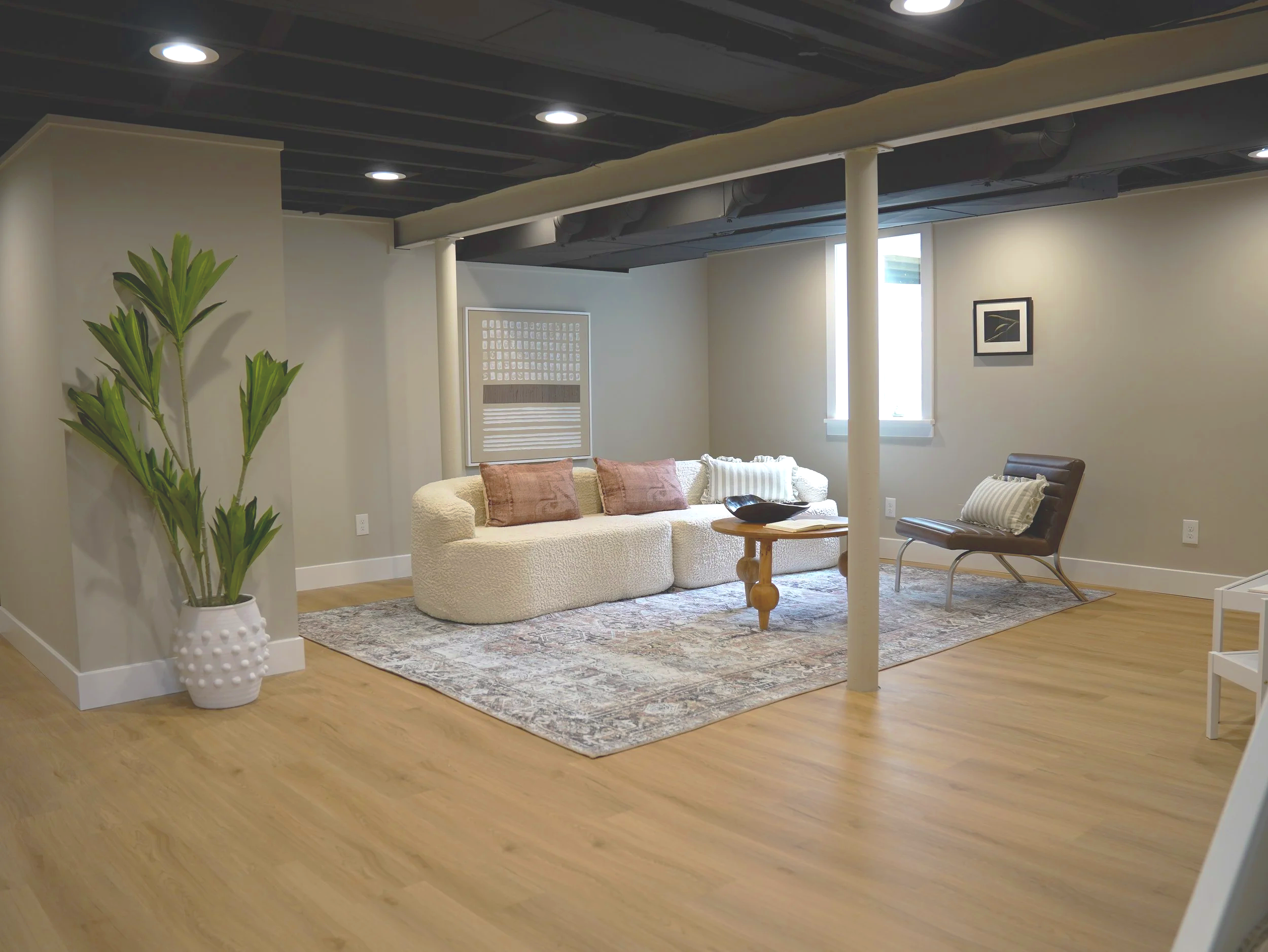 Living room with a beige sofa, pink and white pillows, a small wooden coffee table, brown armchair, a framed picture on the wall, and a window with blinds, with a large potted plant in the foreground, in a room with light hardwood floors and white wa
