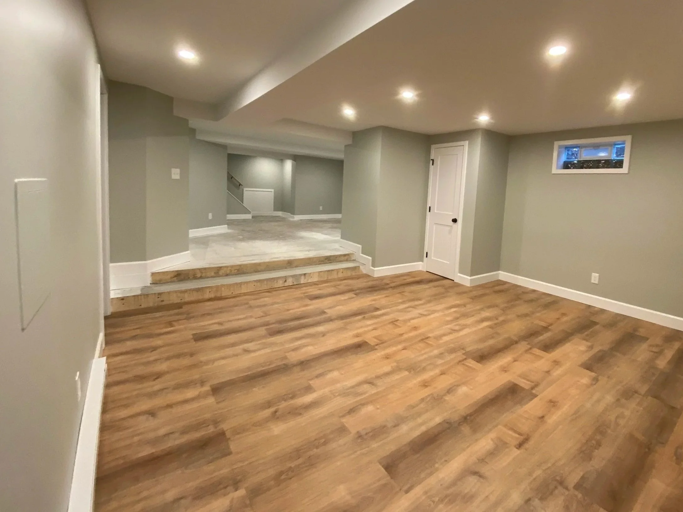 Empty basement room with wood flooring, gray walls, recessed ceiling lights, and a small window.