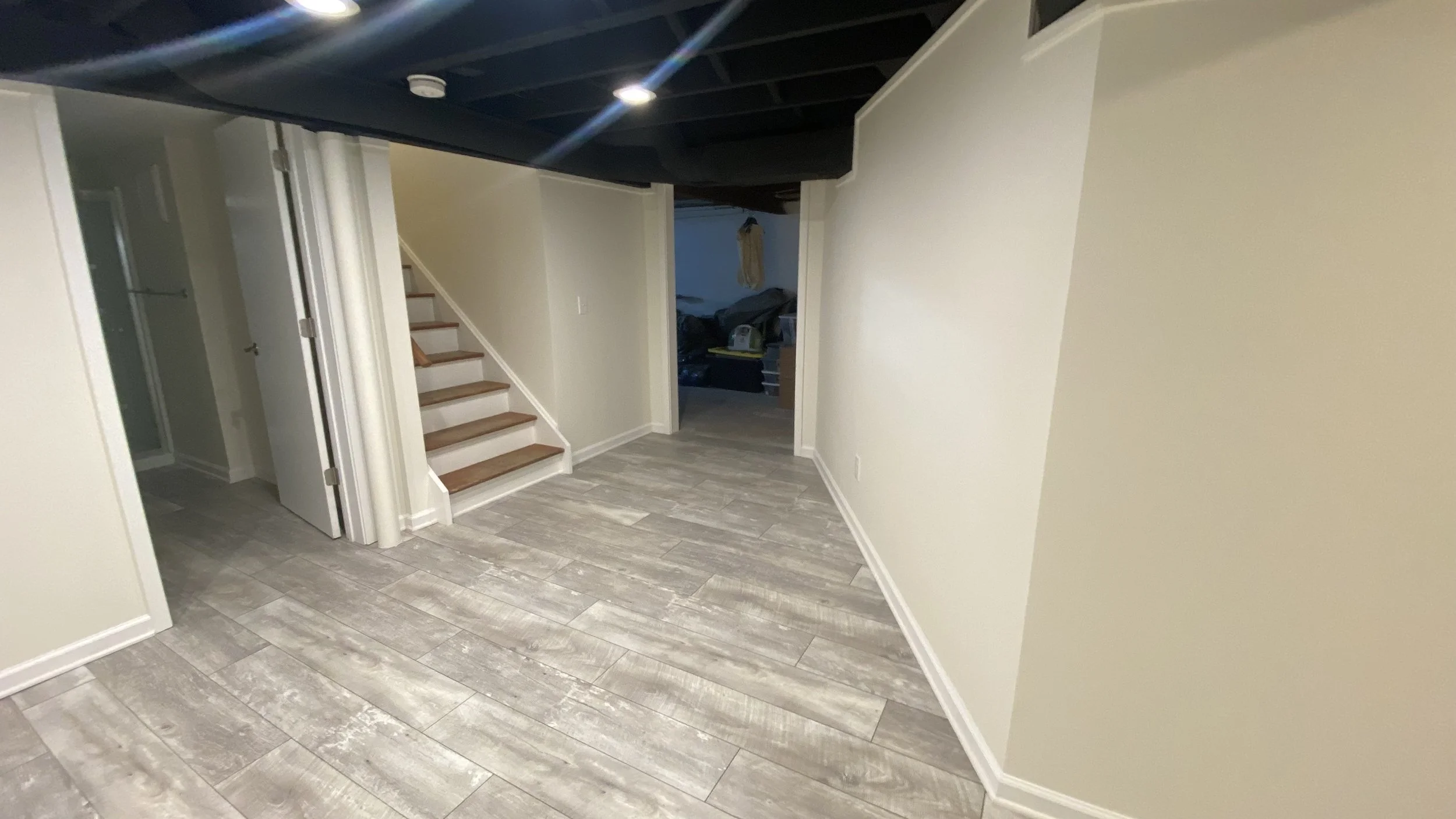 Empty hallway with light gray wood-look tile floors, light-colored walls, stairs with wooden treads and white risers, and a doorway leading to a room with clutter.
