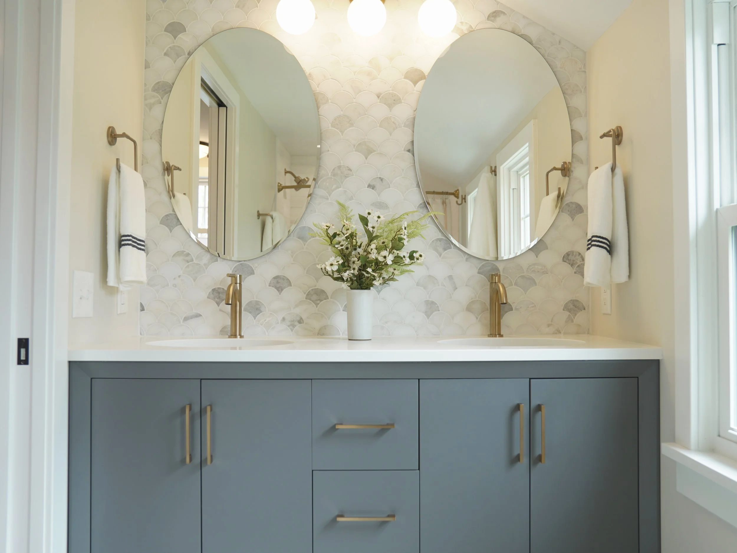 Bathroom vanity with a blue cabinet, gold handles, two mirrors above the sink, a vase with white flowers, and a patterned tile wall. Towel hooks with towels hang on either side of the mirrors.