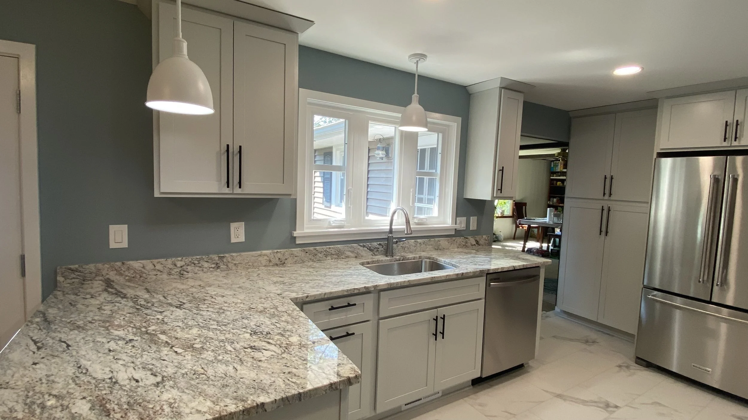 A modern kitchen with white cabinets, granite countertops, a stainless steel sink under a window, and stainless steel appliances, including a refrigerator.
