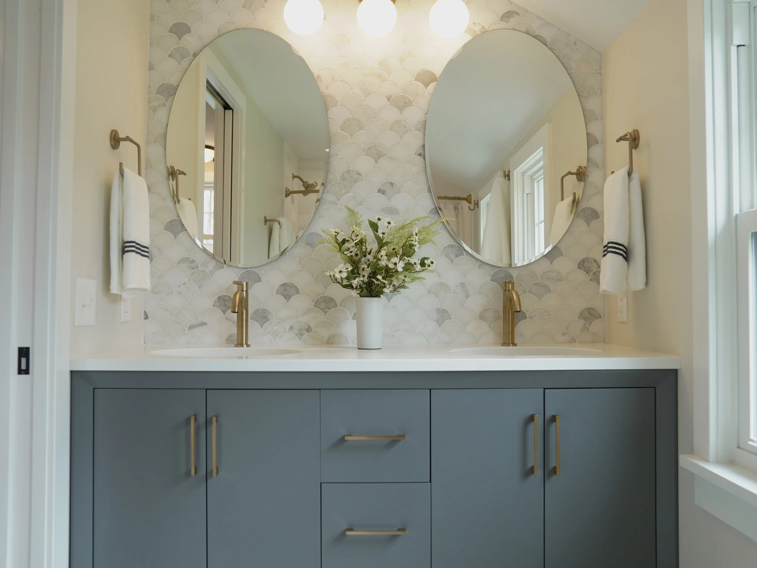 Modern bathroom vanity with navy blue cabinets, gold handles, a white countertop, and a vase with white flowers. Two oval mirrors are mounted above, with wall-mounted gold towel rings holding white towels on either side. A window is on the right, and decorative tiles are on the wall behind the mirrors.