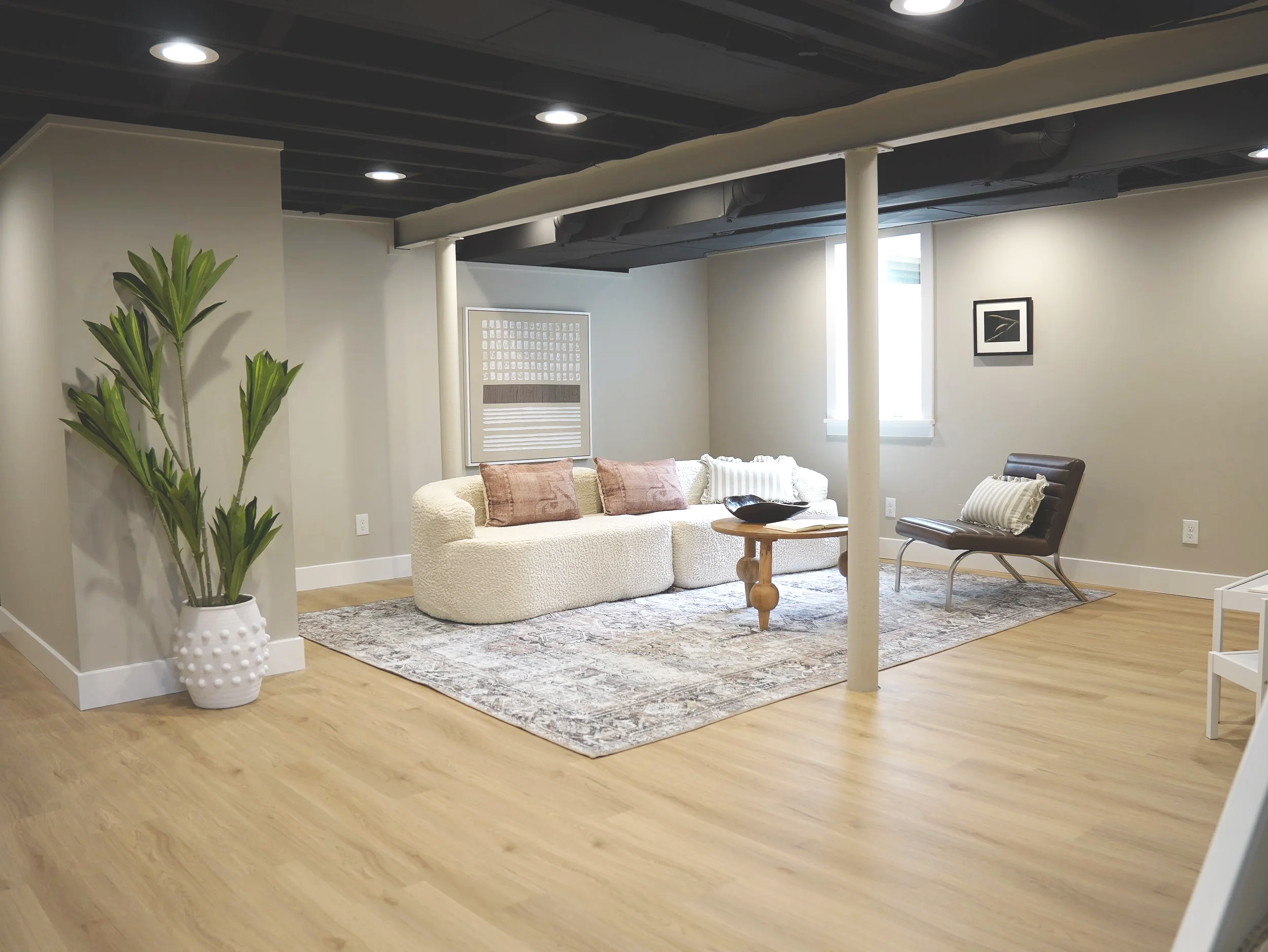 A modern living room with a cream curved sofa, pink and white pillows, a small wooden coffee table, a black leather chair, a framed black-and-white photograph on the wall, a large potted green plant, and a beige and gray area rug on light hardwood floors.