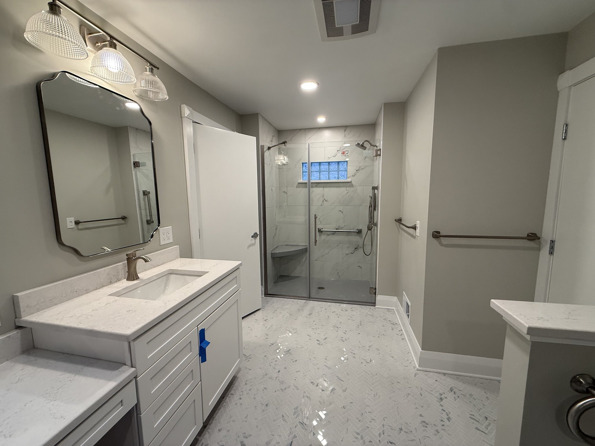 Modern bathroom with a white vanity, marble countertop, and a large mirror, adjacent to a walk-in shower with glass doors and marble tiles, and multiple ceiling lights.