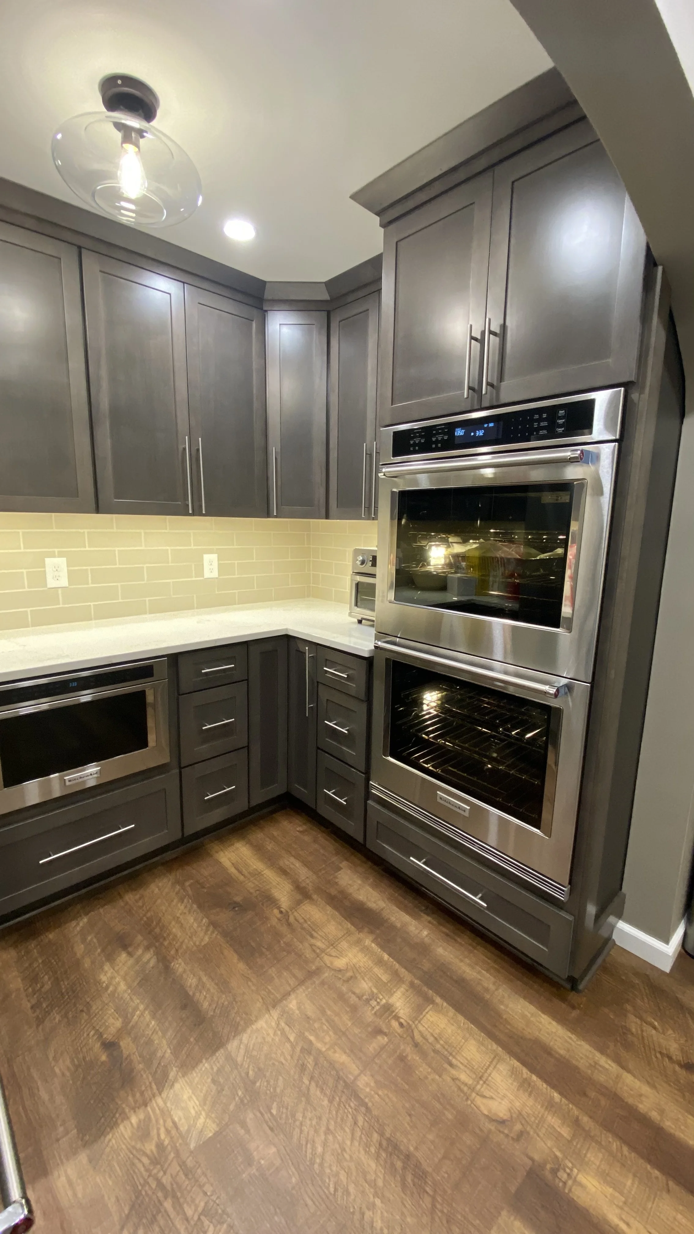 Modern kitchen with dark gray cabinets, stainless steel oven and microwave, beige subway tile backsplash, white countertops, and wooden flooring.