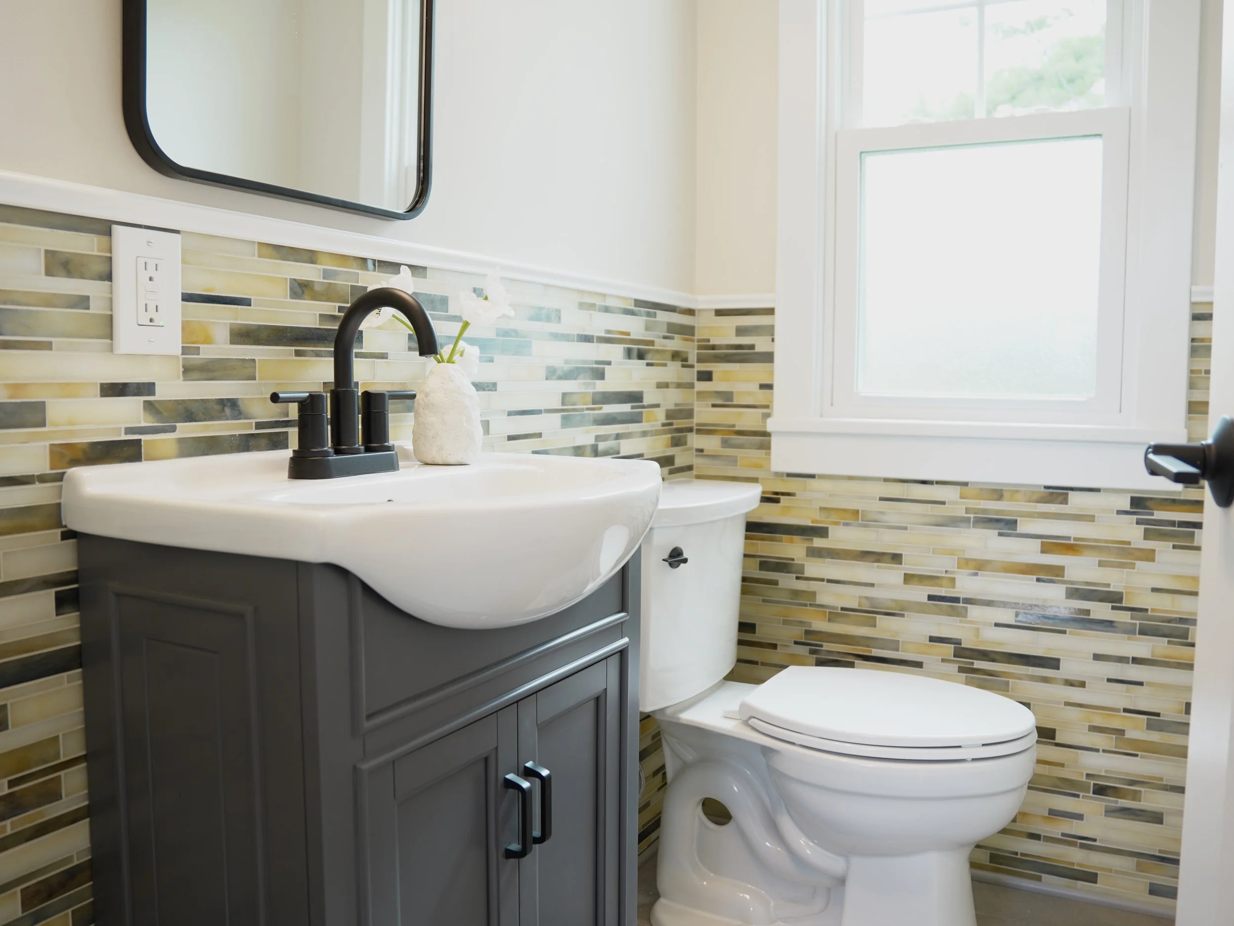 A modern bathroom with a black faucet and white sink, a decorative vase with white flowers, a tiled backsplash, a window letting in natural light, and a white toilet.