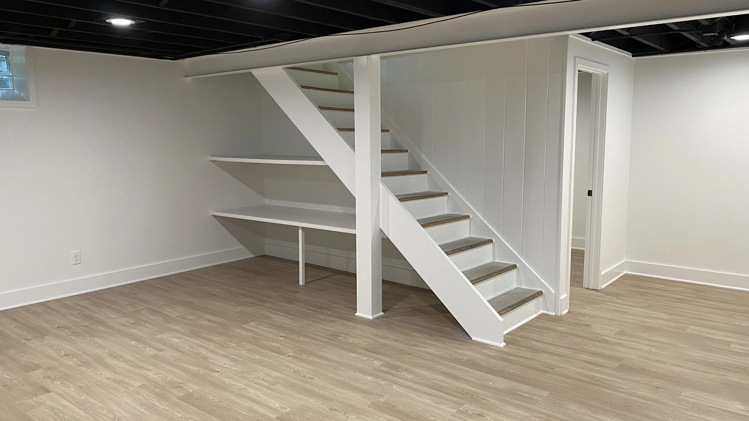 Interior of a house with a white staircase, light-colored wooden flooring, white walls, and partially exposed ceiling with black beams.