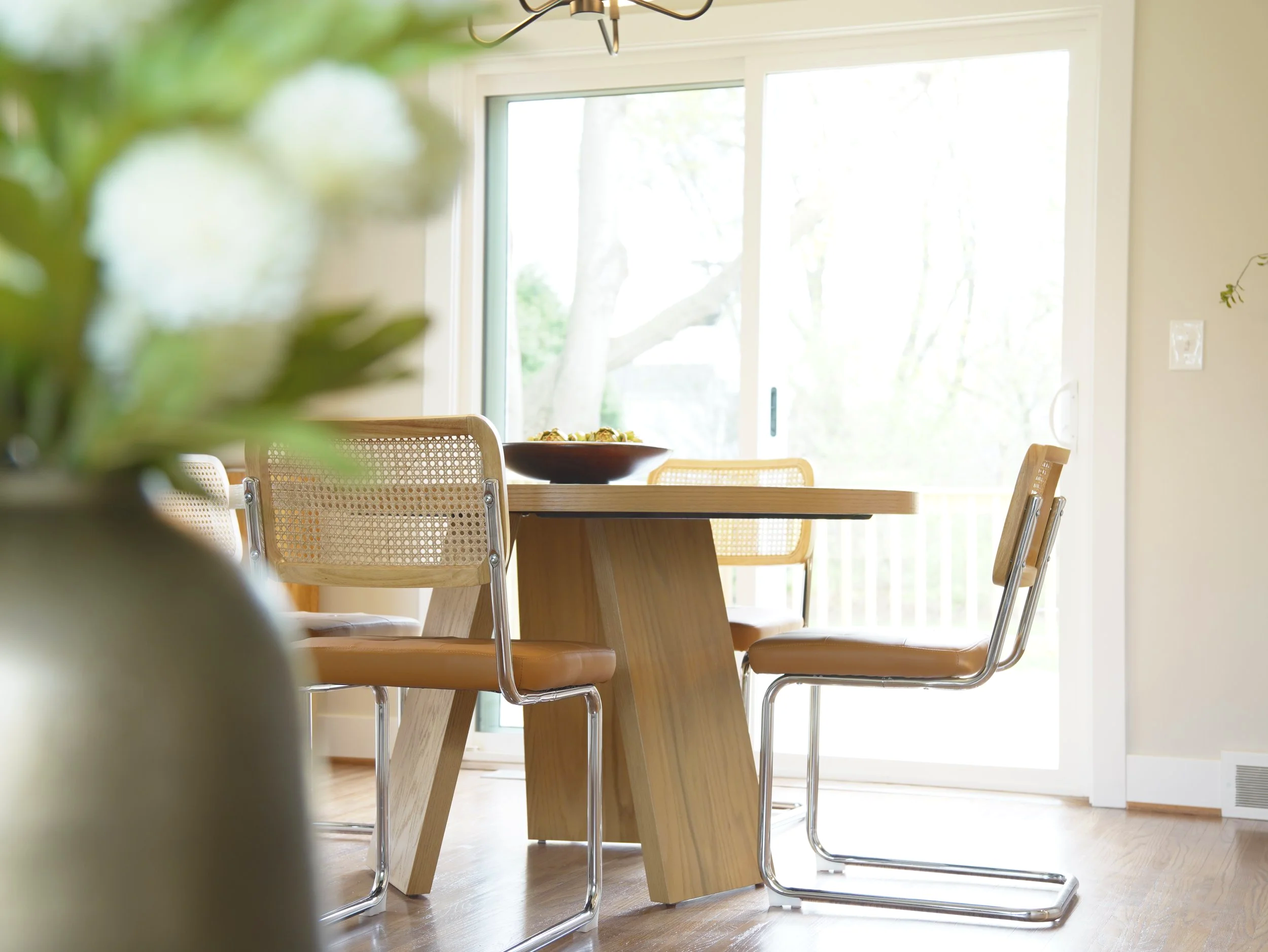 A wooden dining table with beige chairs around it, a bowl of food, large windows with trees outside, a ceiling light fixture, and a light-colored wall with an electrical outlet.
