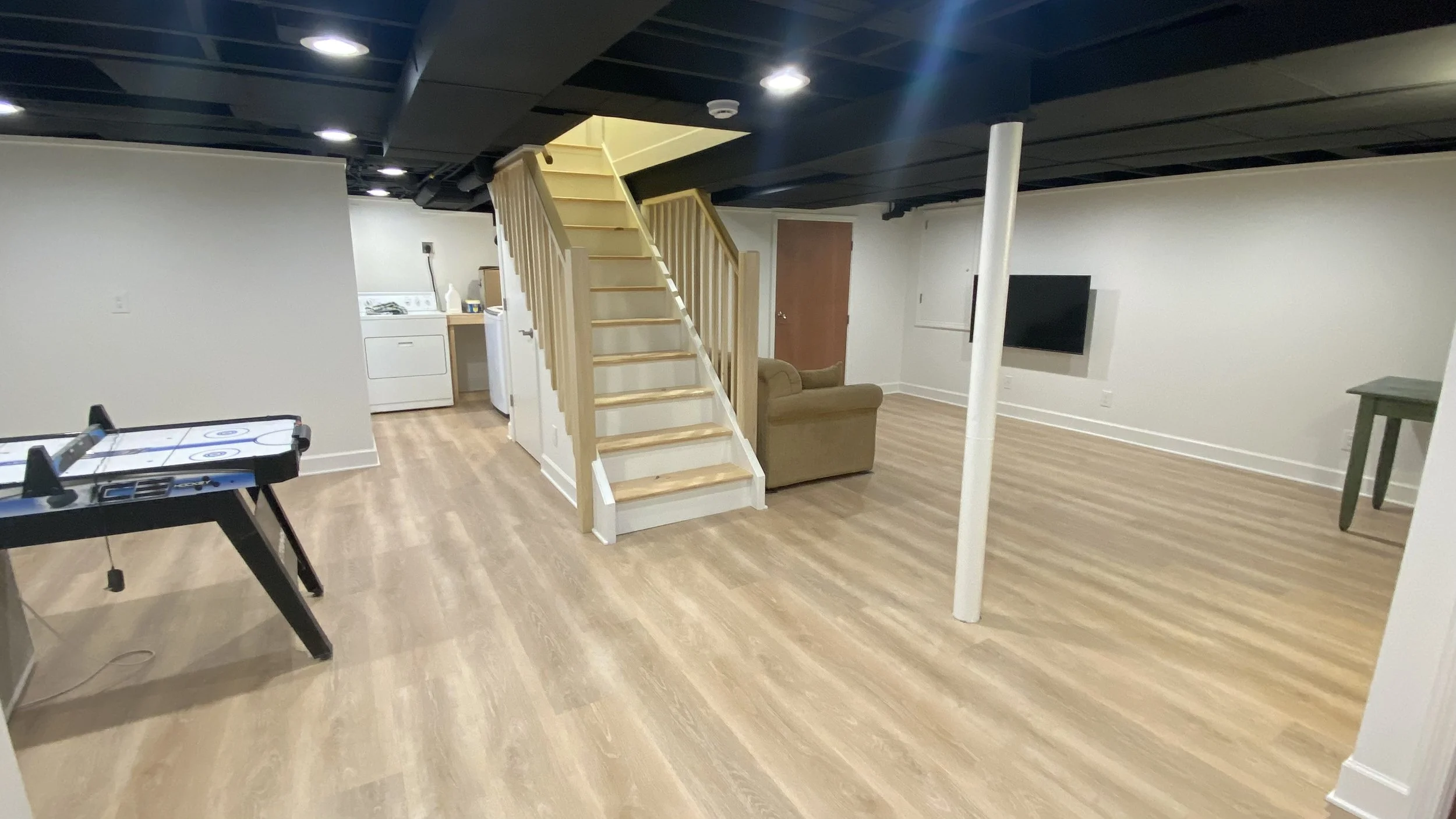 Basement room with wooden stairs, a couch, mounted TV, laundry area, air hockey table, and a ping pong table.