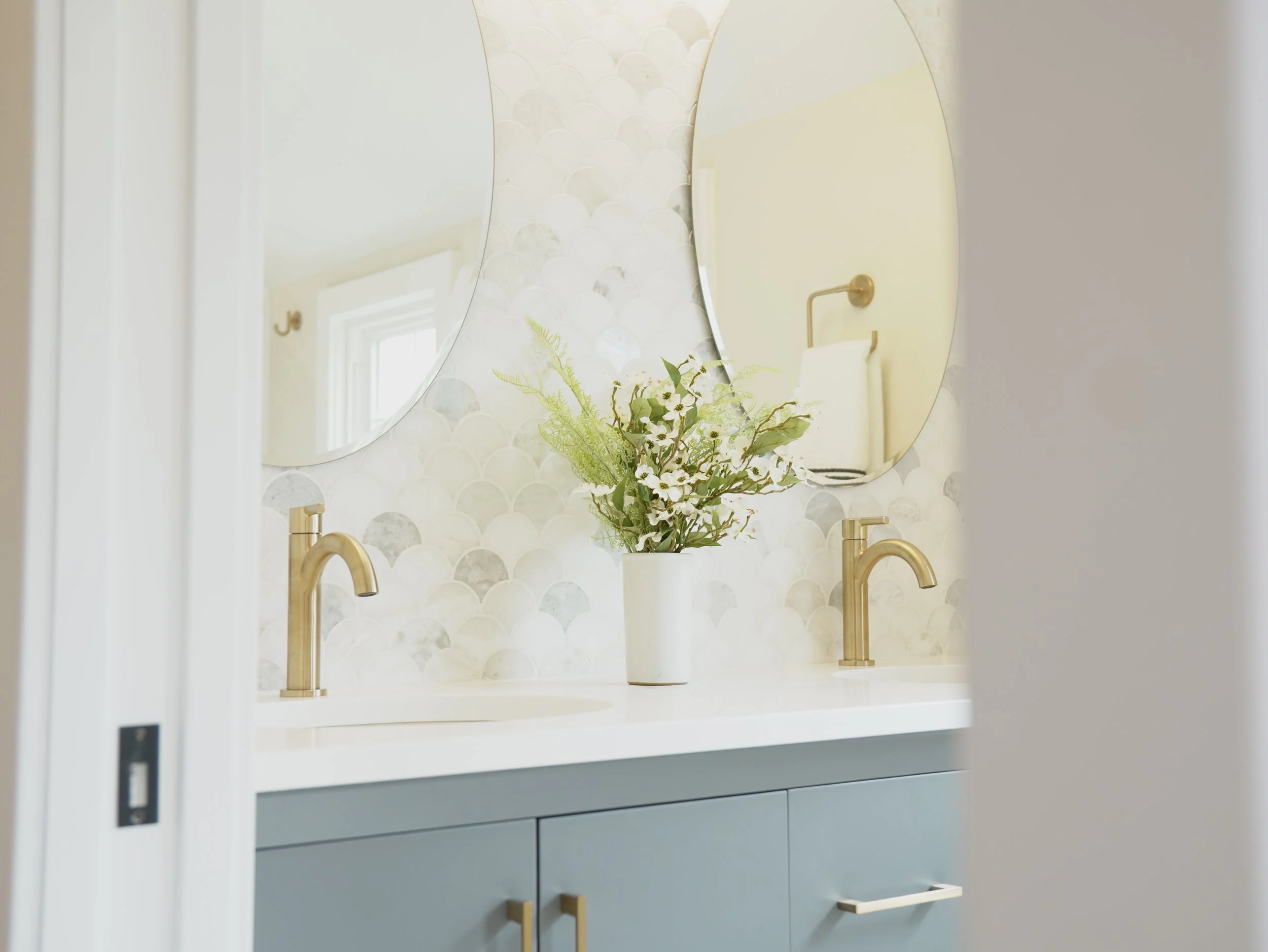 A modern bathroom vanity with a white countertop, gold faucet, a potted plant with greenery and white flowers, and two oval mirrors mounted on a tiled wall with a scalloped pattern.