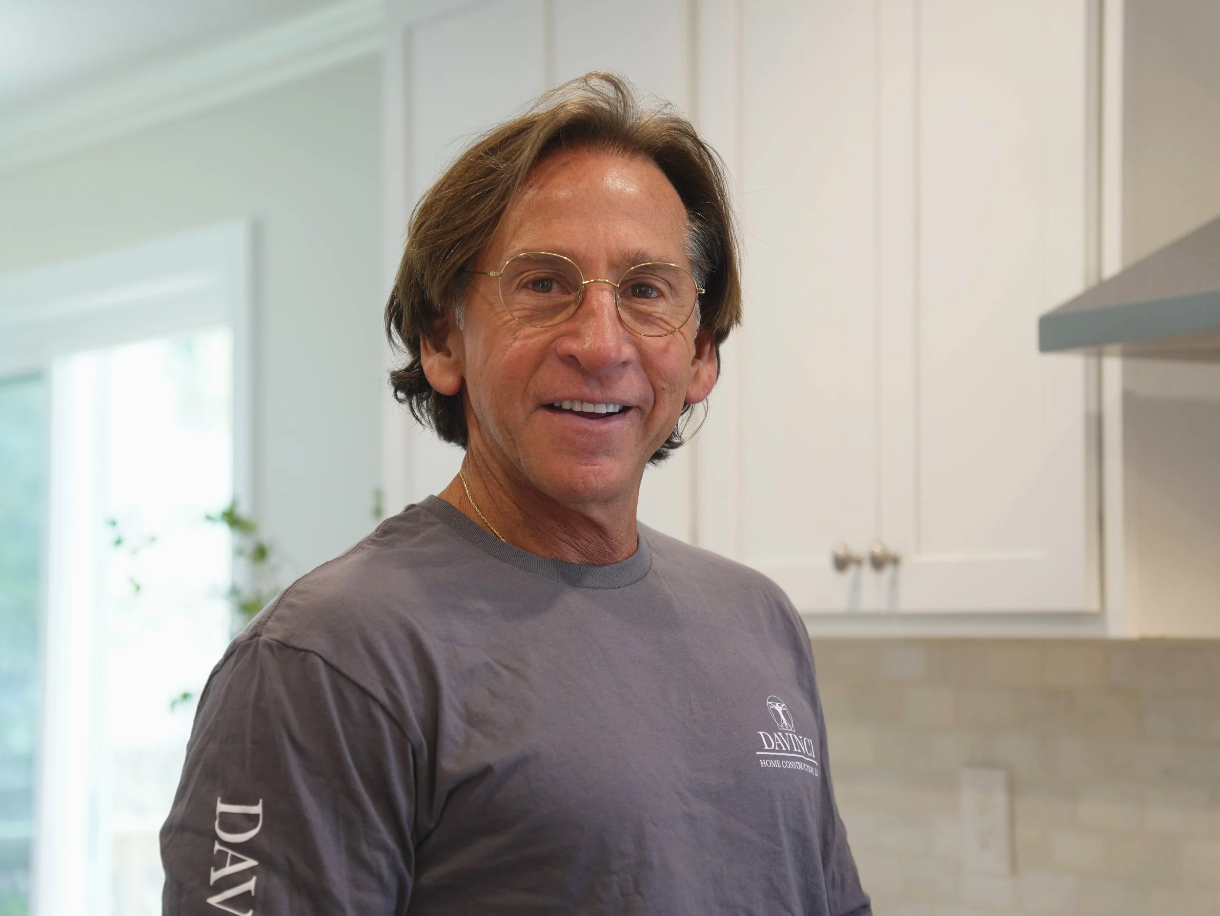 A middle-aged man wearing glasses and a gray t-shirt with a logo, smiling in a bright kitchen with white cabinets and a window.