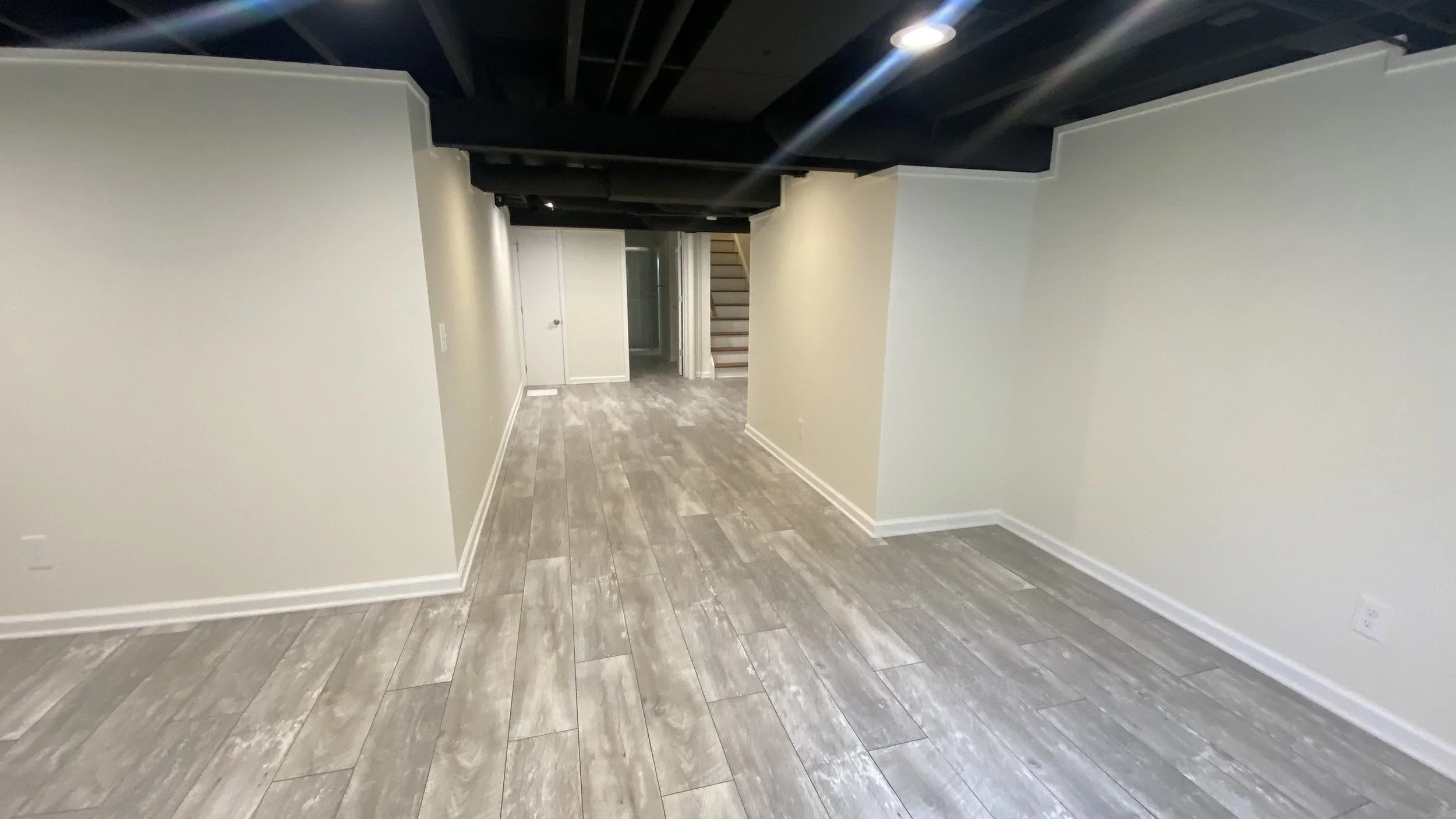 An empty basement with light gray wood flooring, beige walls, and white trim. There are stairs leading up at the back, with a closed white door at the start. A black ceiling with visible beams and a round ceiling light fixture is overhead.