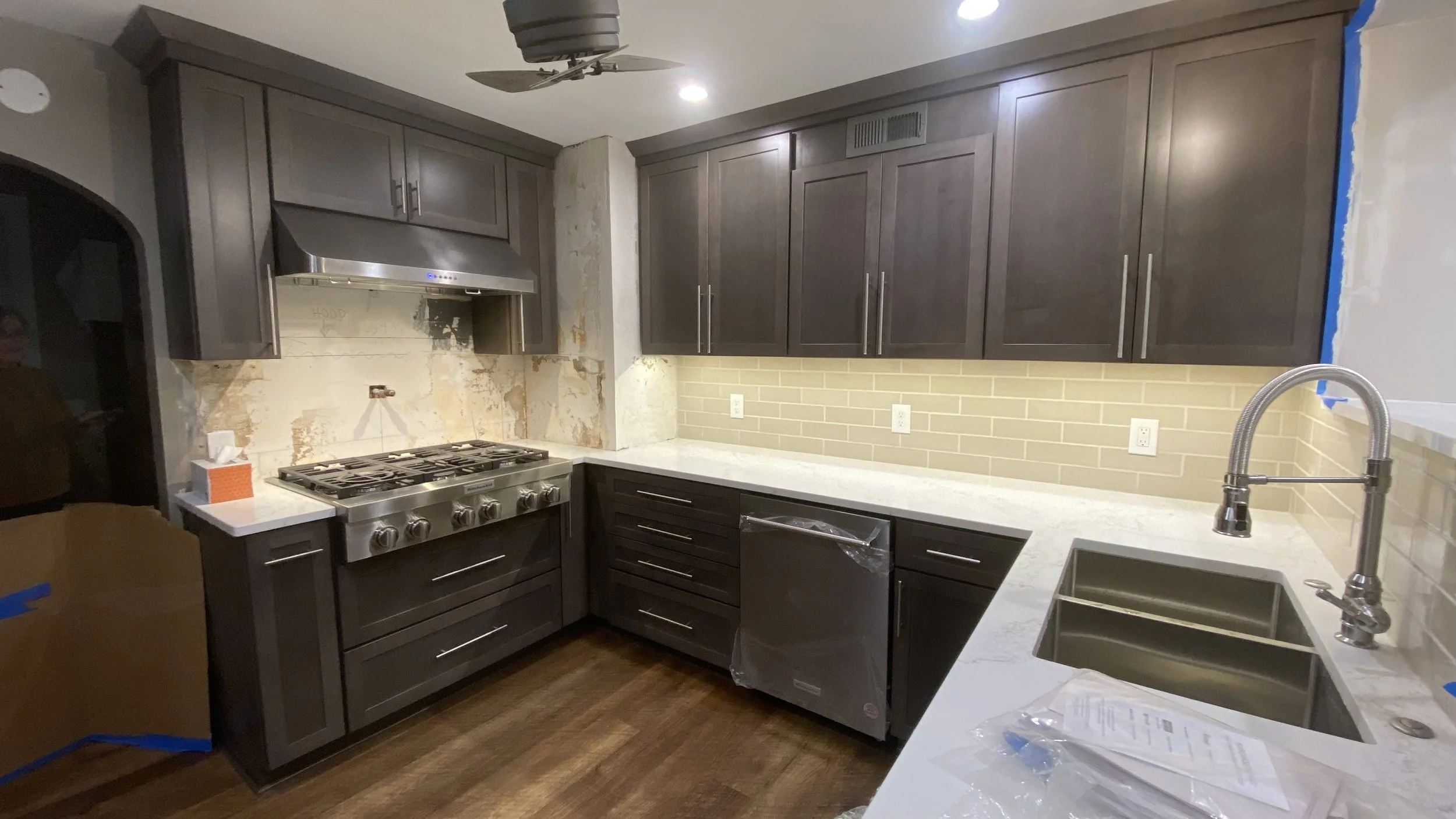 Kitchen with dark cabinets, white countertops, a stainless steel stove and hood, a double sink, a yellow brick backsplash, and ongoing construction with masking tape on the walls.