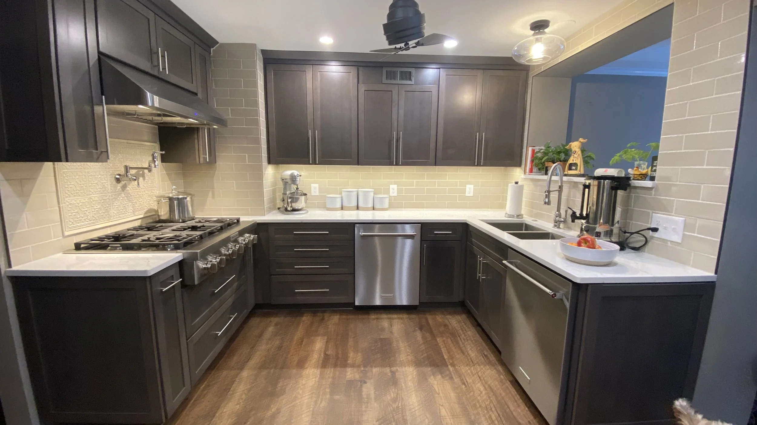 A modern kitchen with dark gray cabinets, beige subway tile backsplash, and stainless steel appliances, including a stove, dishwasher, and microwave. The kitchen has a white countertop, a double sink with a faucet, a paper towel holder, and various a