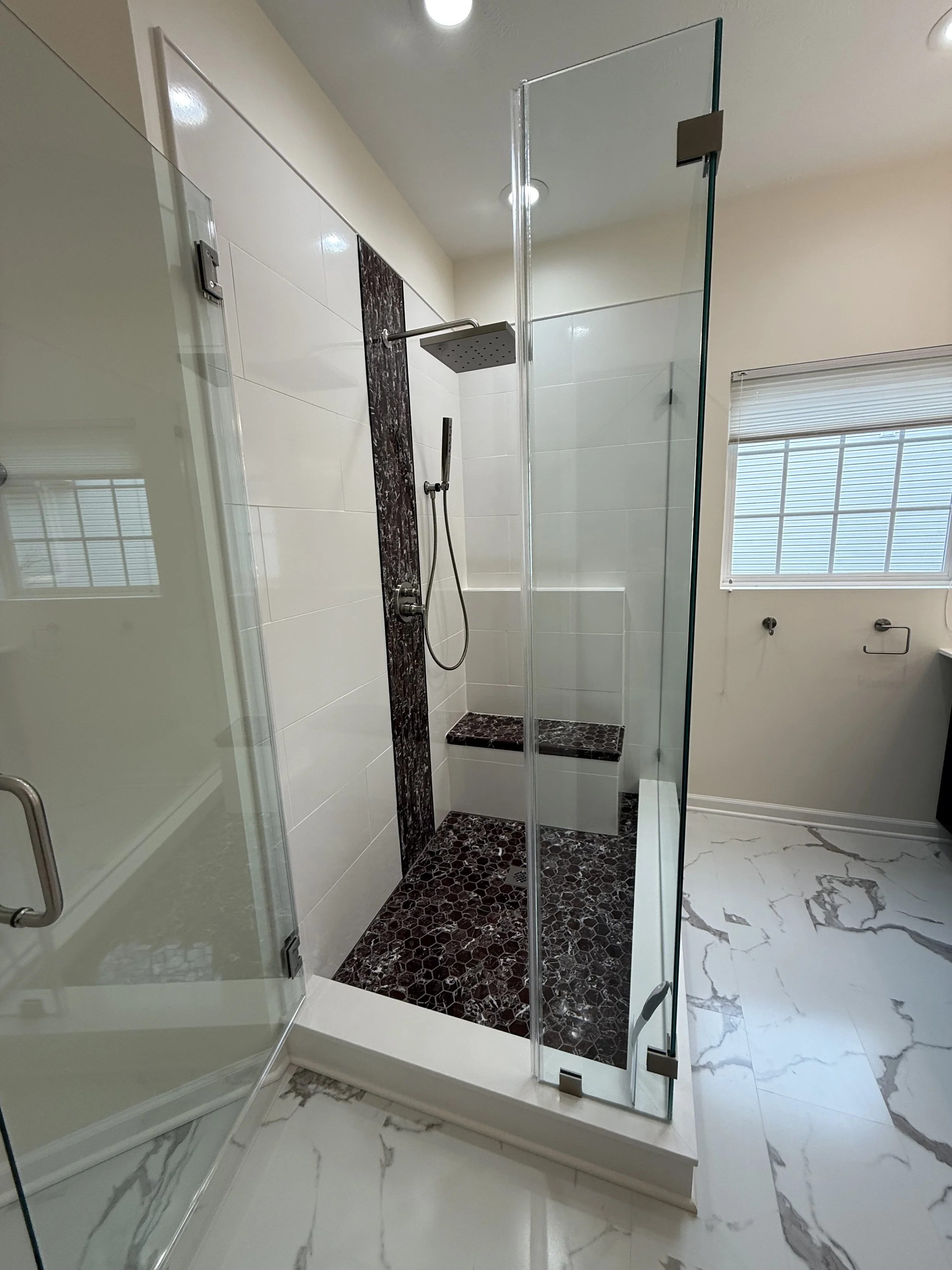 A modern walk-in shower with a glass enclosure, rainfall showerhead, and stone tiles. The shower has a dark pebble floor and a vertical dark marble stripe on the wall. There's a window with blinds nearby and marble flooring outside the shower.
