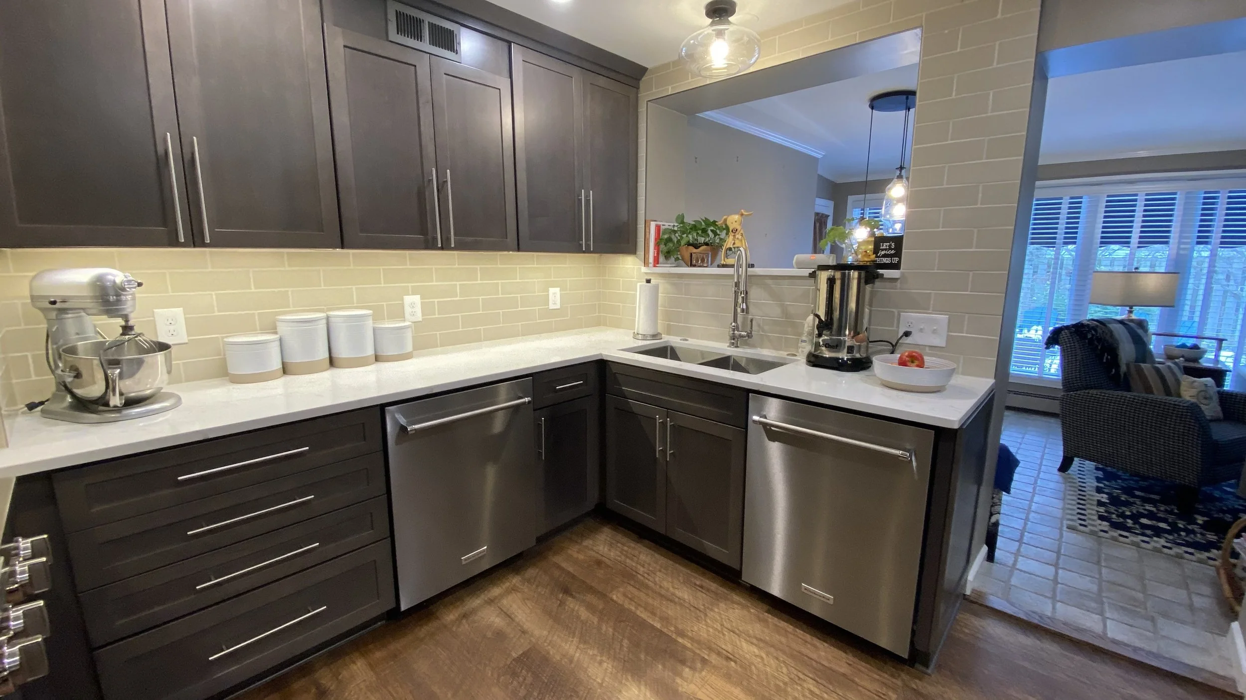 Modern kitchen with dark gray cabinets, white countertop, and beige brick backsplash. Contains a stand mixer, three white canisters, a paper towel holder, and a blender. Adjacent to the kitchen is a living area with a chair, table, and large window.