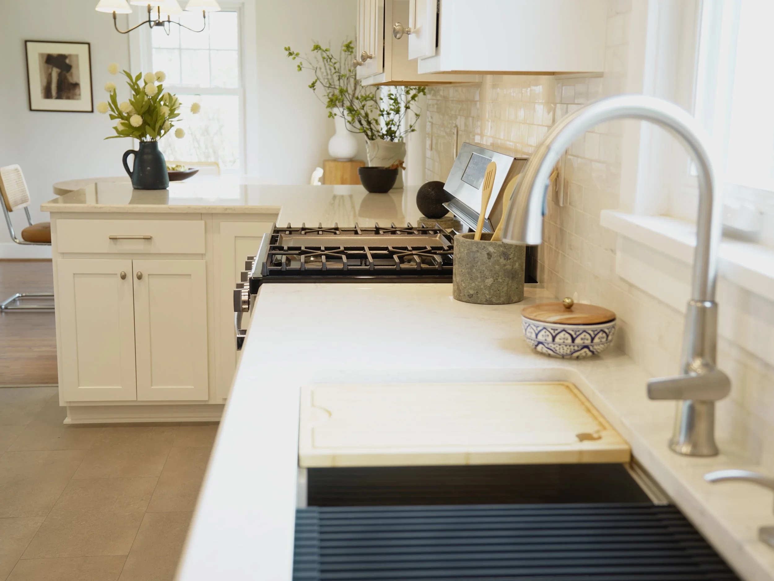 A modern kitchen with white cabinetry, a marble countertop, a gas stove, and decor including vases with flowers. There is a window in the background allowing natural light into the space.